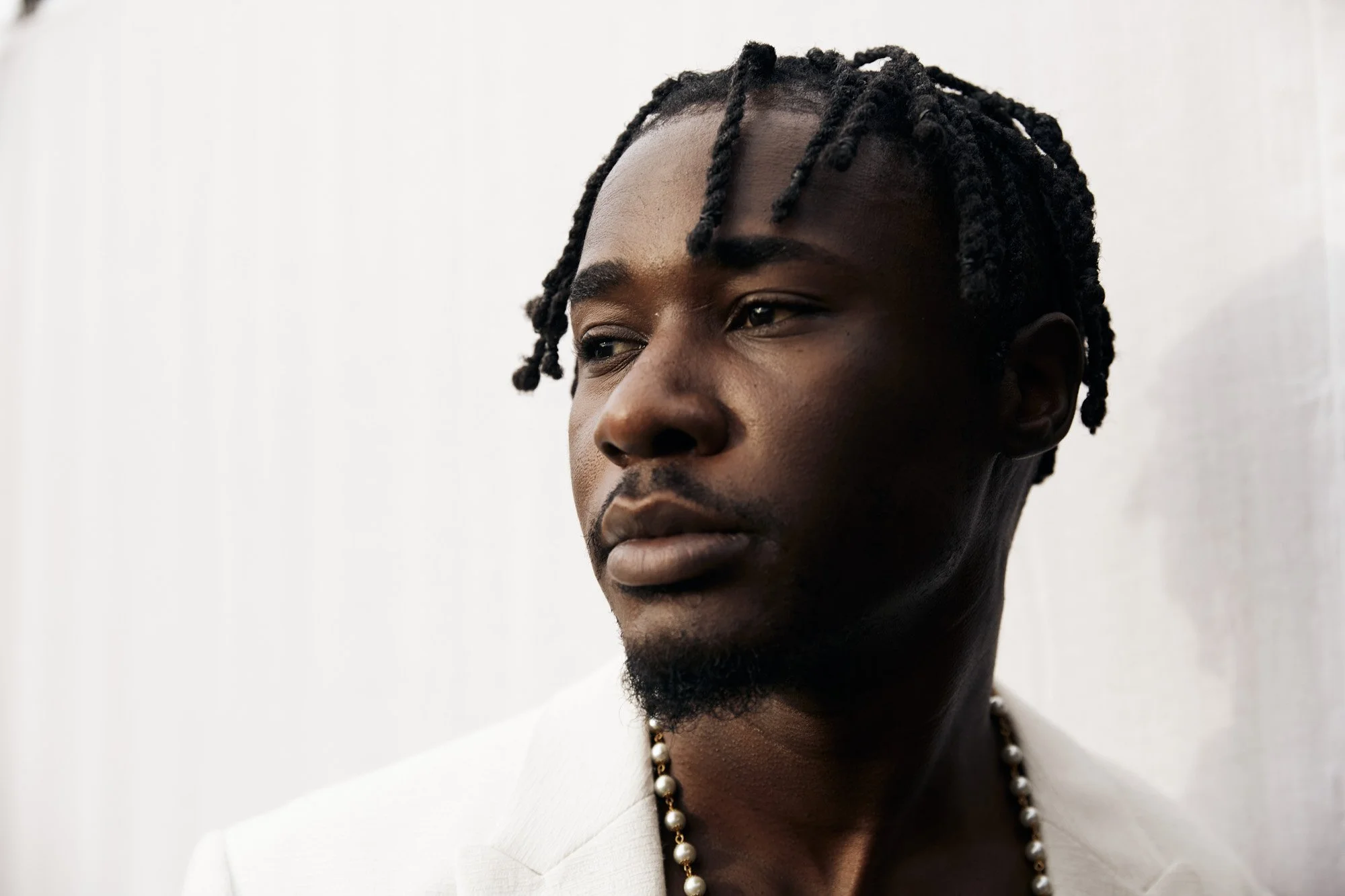 Portrait of a young Black man with styled twists in his hair, wearing a white blazer and a pearl necklace, looking to the side with a neutral expression.