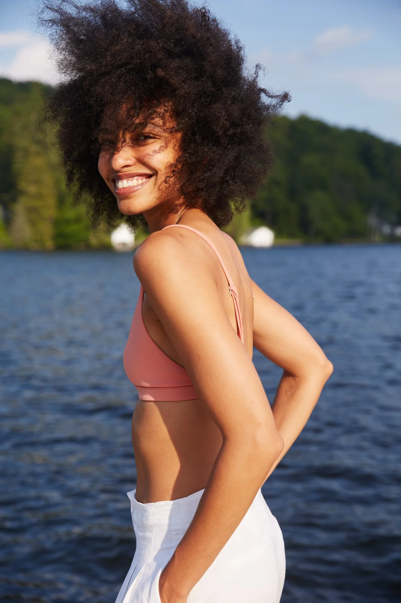 Avril Guerrero with curly hair smiling by a lake during the day, wearing a pink top and white pants.