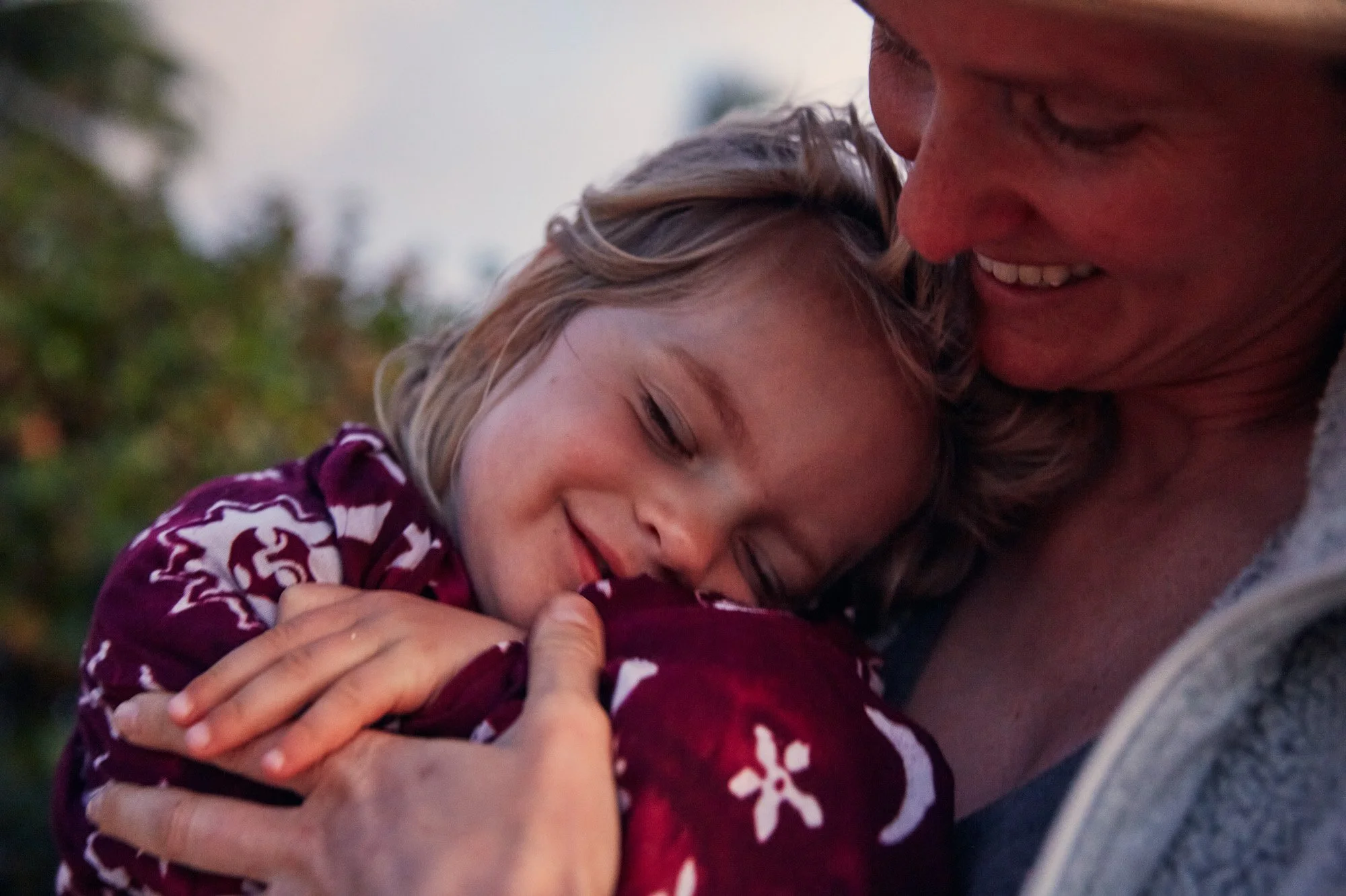 A young girl with blonde hair smiling and hugging her mother tightly, both sharing a tender moment outdoors.