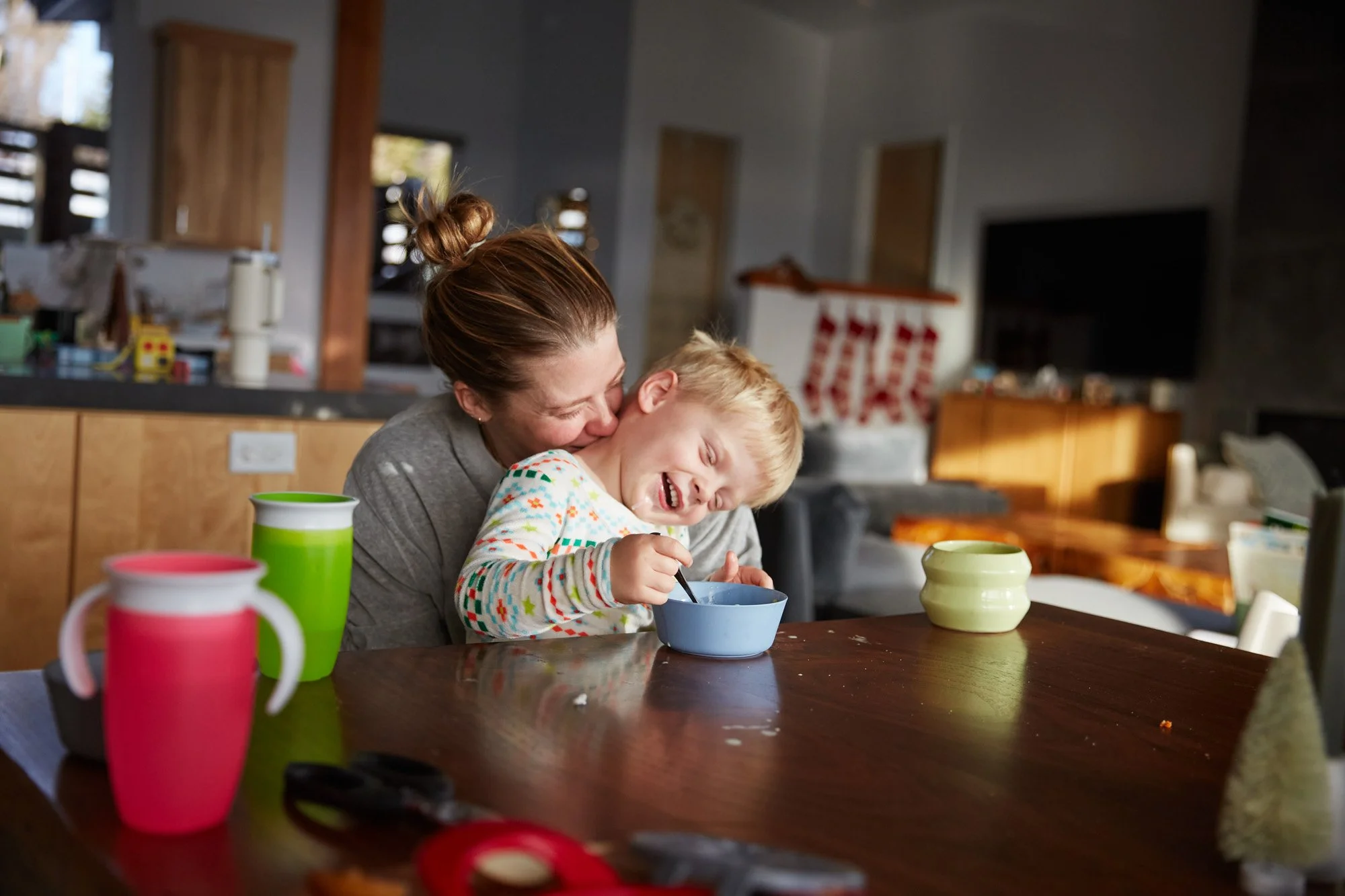 A woman playfully hugging her laughing young boy at a kitchen table during breakfast, with colorful cups and a bowl.