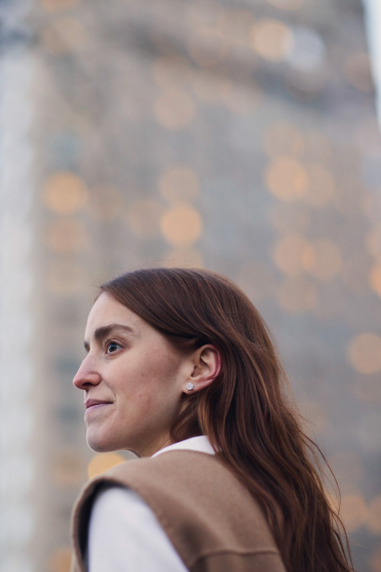 Profile of Ana Corina Sosa with long brown hair, wearing earrings, and a brown coat, with a blurred cityscape in the background. Central Park Manhattan New York City