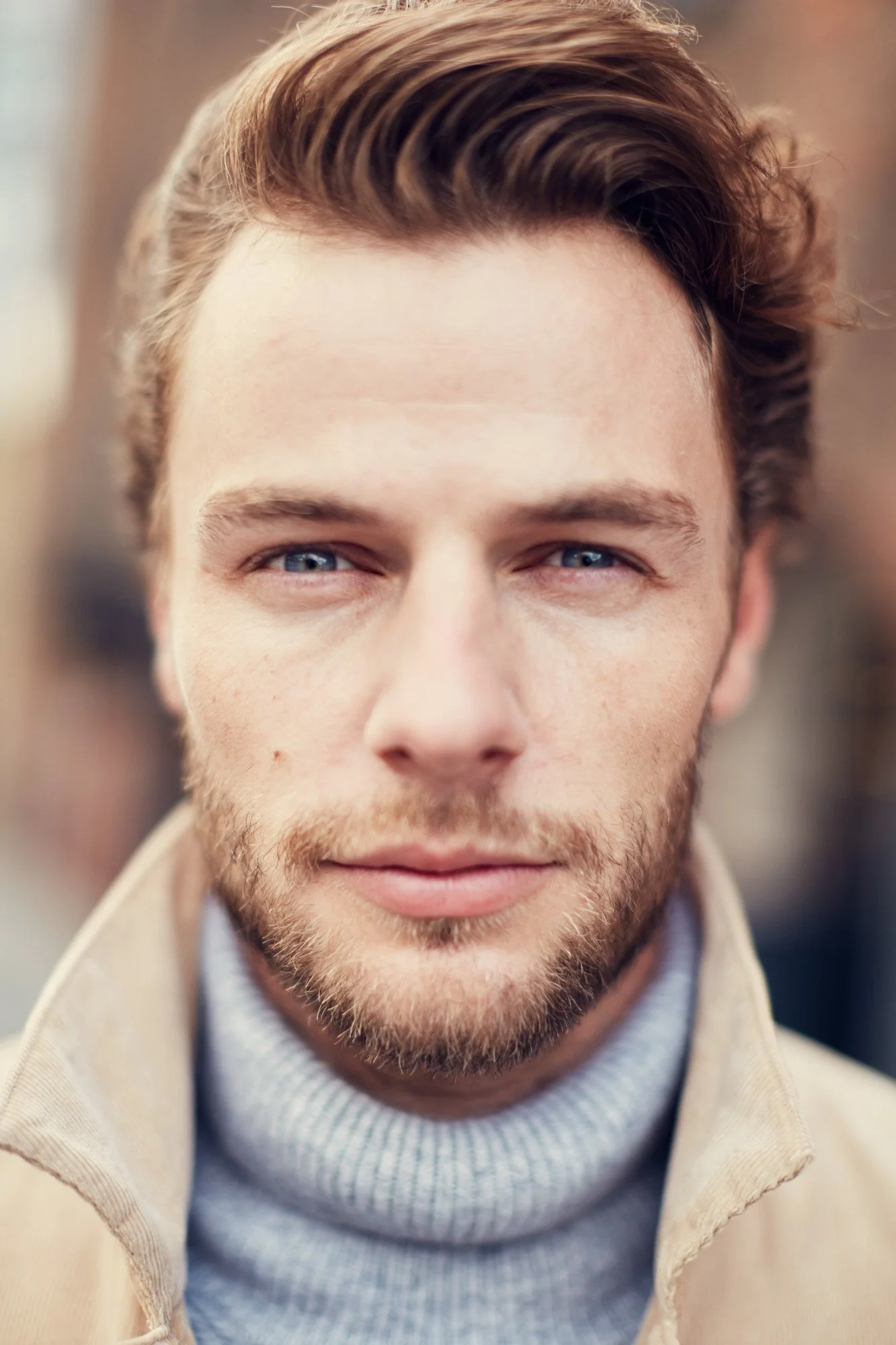 Close-up portrait of a young man with light brown hair, blue eyes, and facial hair, wearing a beige jacket over a gray turtleneck sweater.