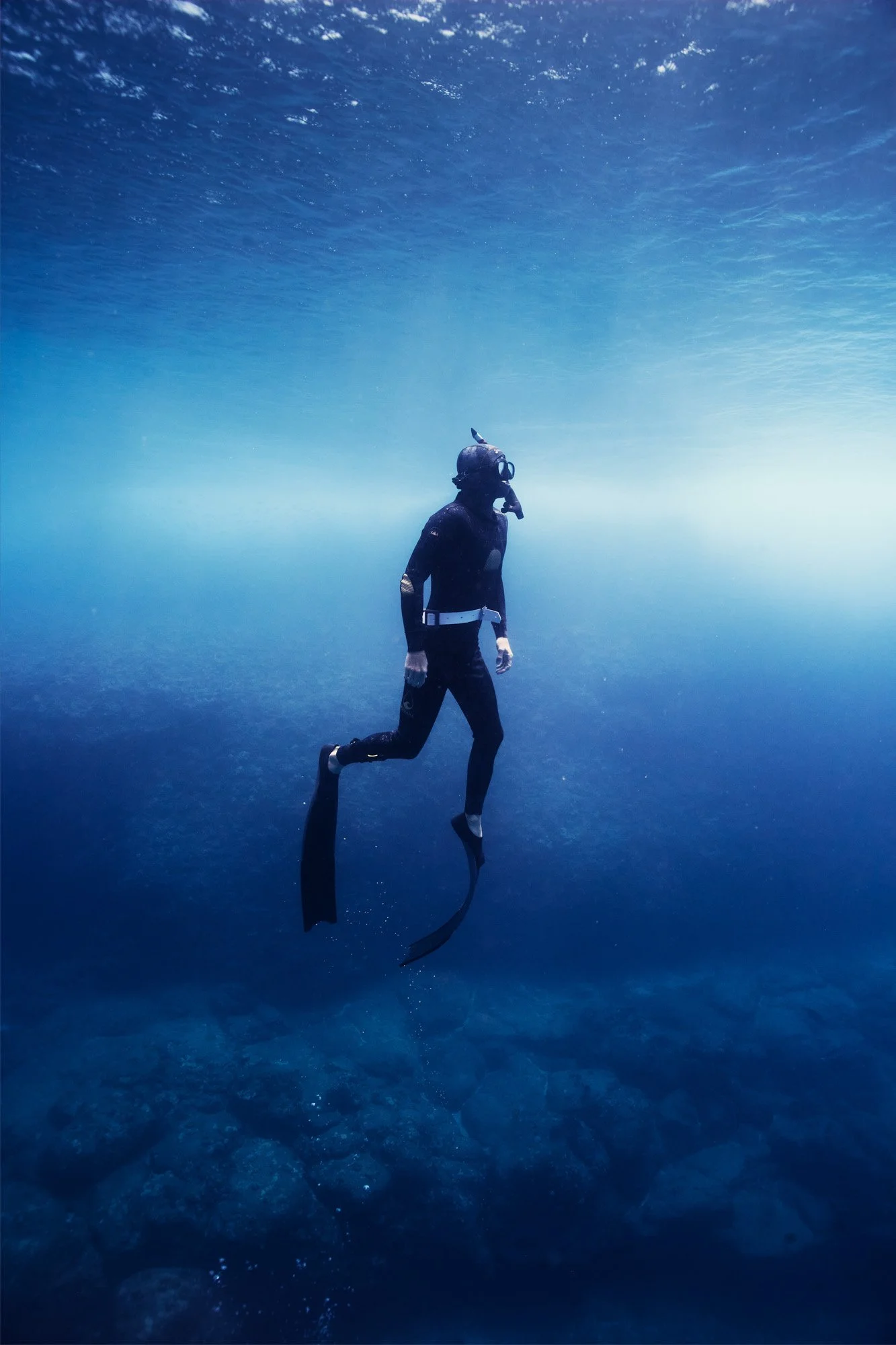 A person scuba diving underwater, wearing a wetsuit, mask, and fins, surrounded by blue water and rocks on the ocean floor. Shark's Cove Oahu Hawaii