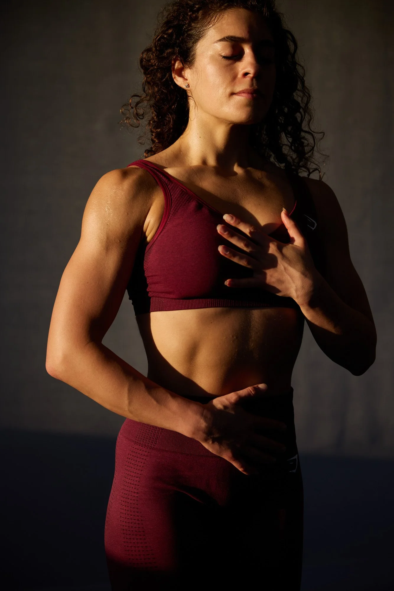 A woman with curly hair wearing a maroon sports bra and matching leggings, practicing yoga or meditation with her eyes closed, one hand on her chest and the other on her abdomen, in a dimly lit environment.