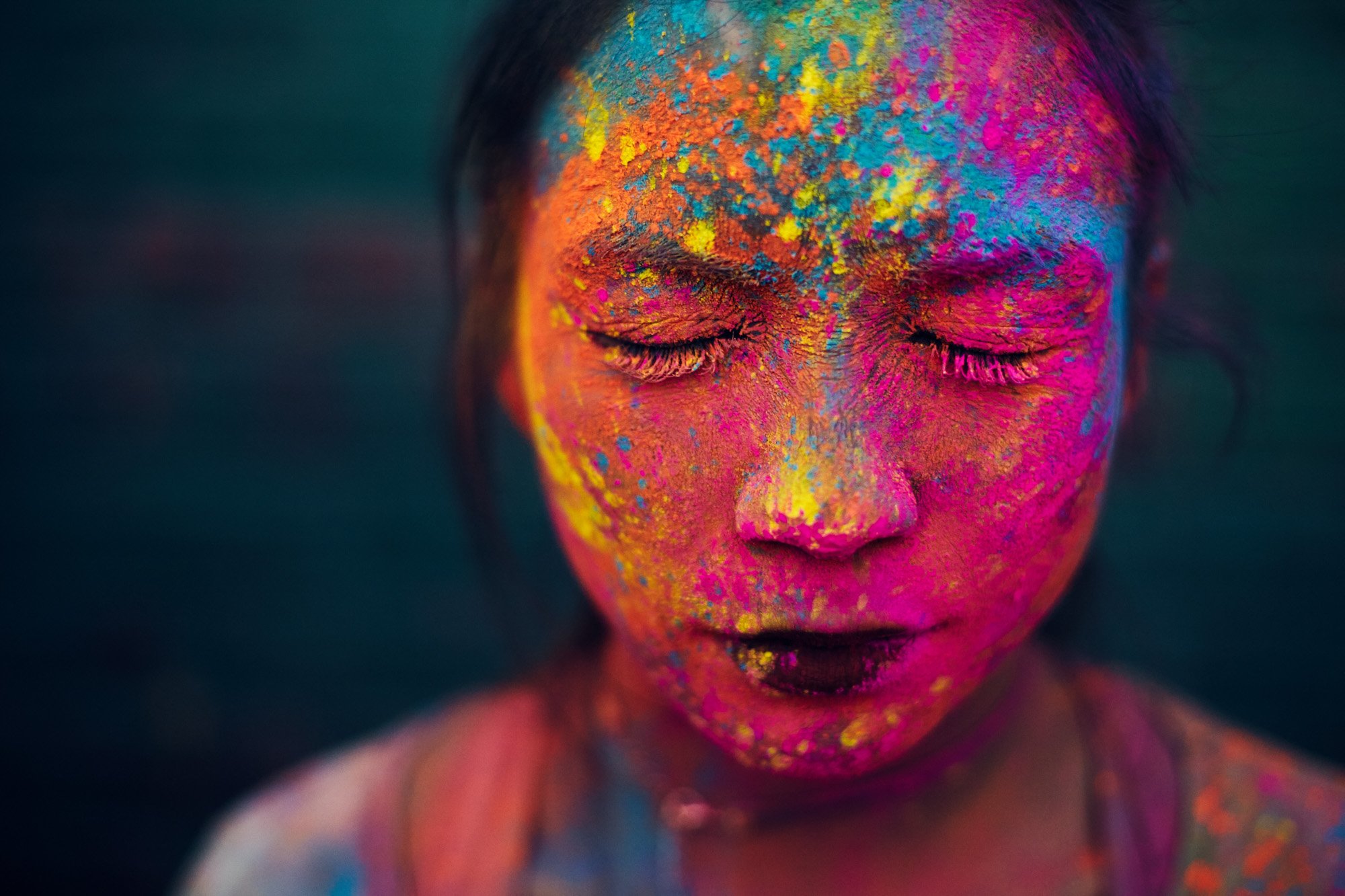 Close-up of a young woman's face with eyes closed, covered in colorful powder in shades of pink, orange, yellow, blue, and purple, during a holi festival.