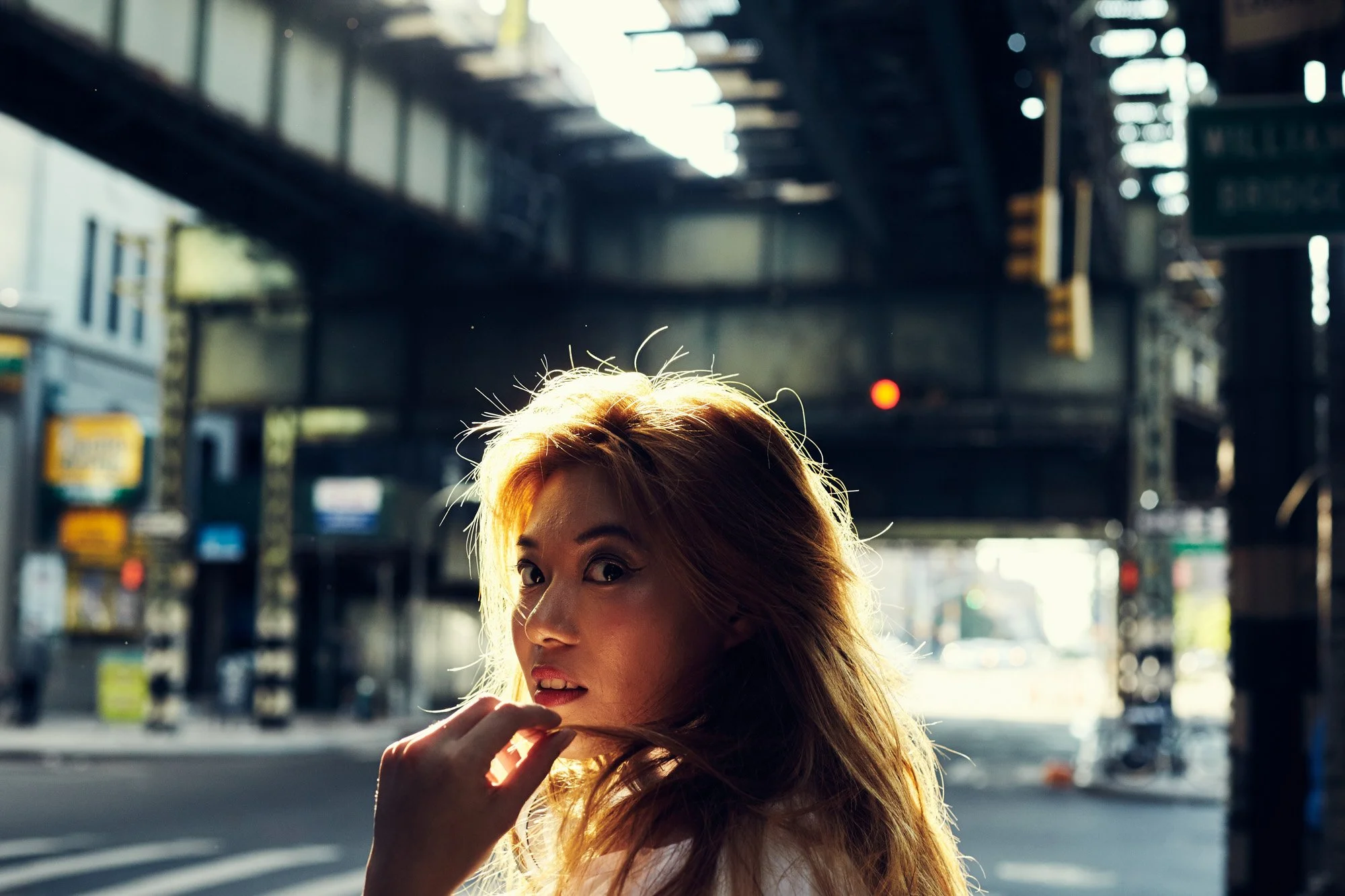 DJ Katsy Lee, with long, light-colored hair standing under an elevated train track in Williamsburg Brooklyn New York City, with sunlight illuminating her face from behind.