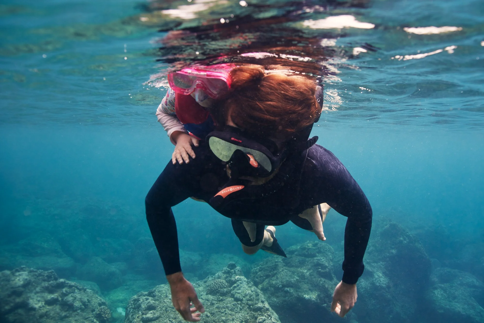 A father snorkeling wearing a black wetsuit and goggles, with a child riding on his back holding onto his shoulders, in a clear ocean with rocks visible below.
