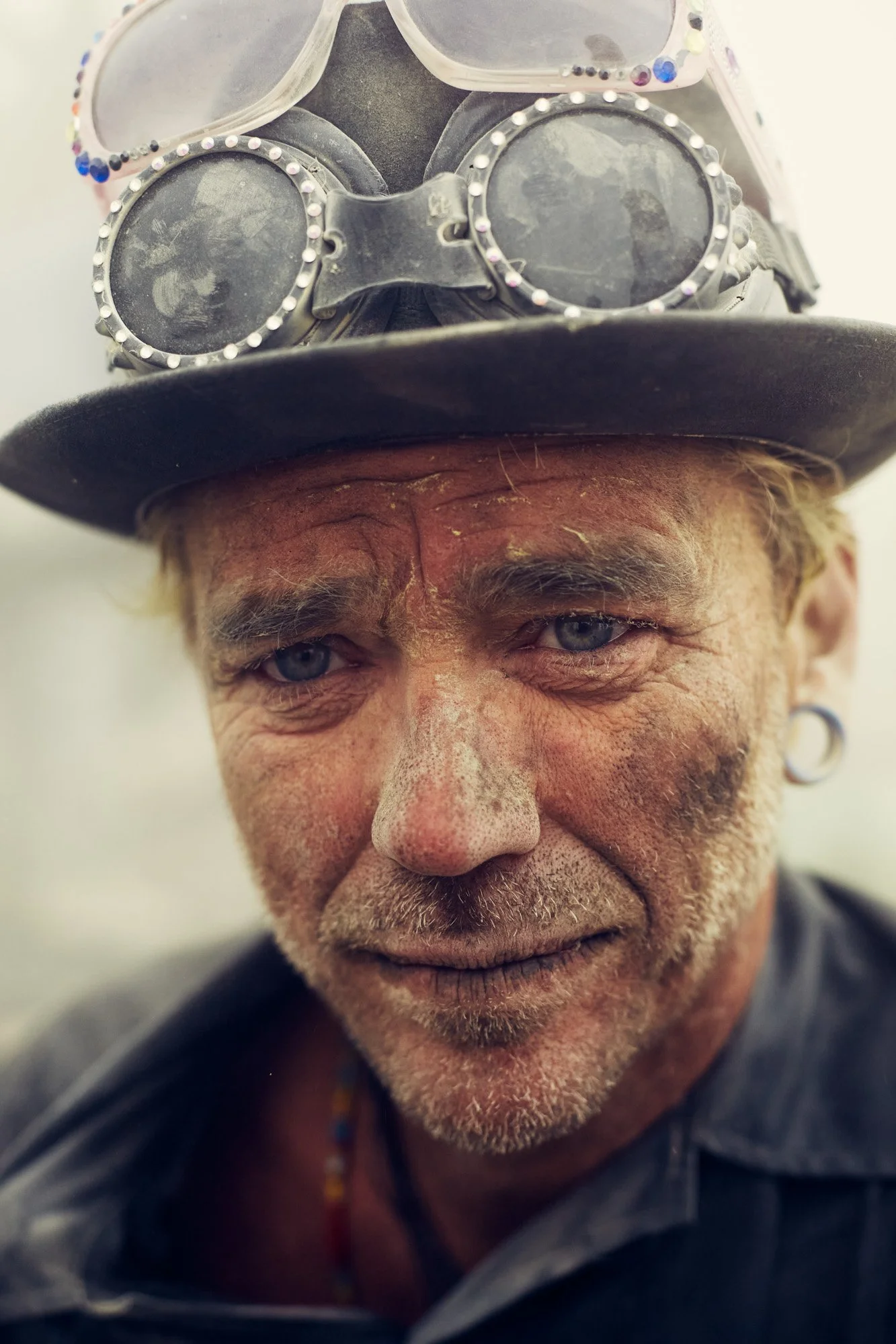 Close-up of a man with dirt-smudged face, wearing a black top hat with goggles on it, and a dark shirt. In the ashes of the burnt man, Burning Man