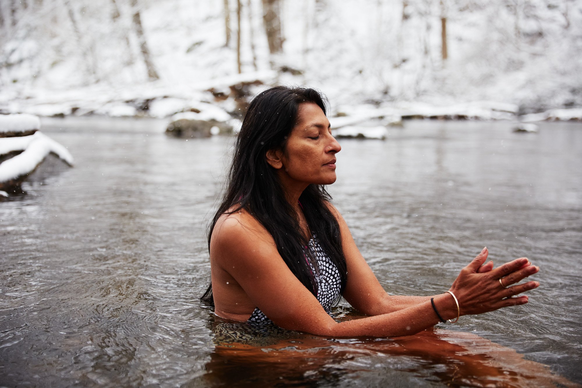 Swati Thiyagarajan, meditating in a river with snow-covered trees and rocks in the background, in Stamford Connecticut, with her eyes closed and hands in prayer position.