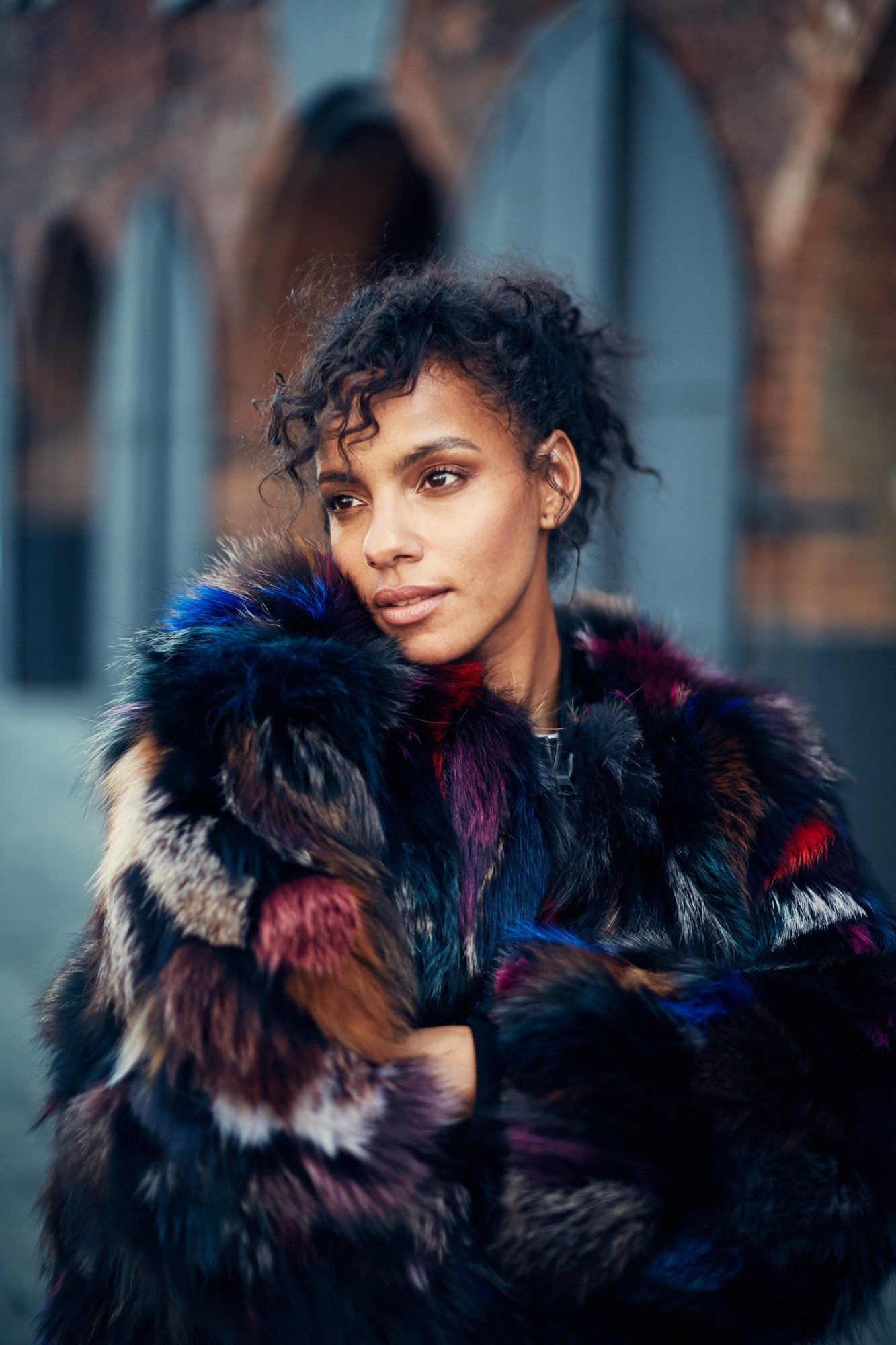 A woman with curly dark hair wearing a multicolored fur coat, standing outdoors in front of a brick building with arched windows. DUMBO Brooklyn New York City