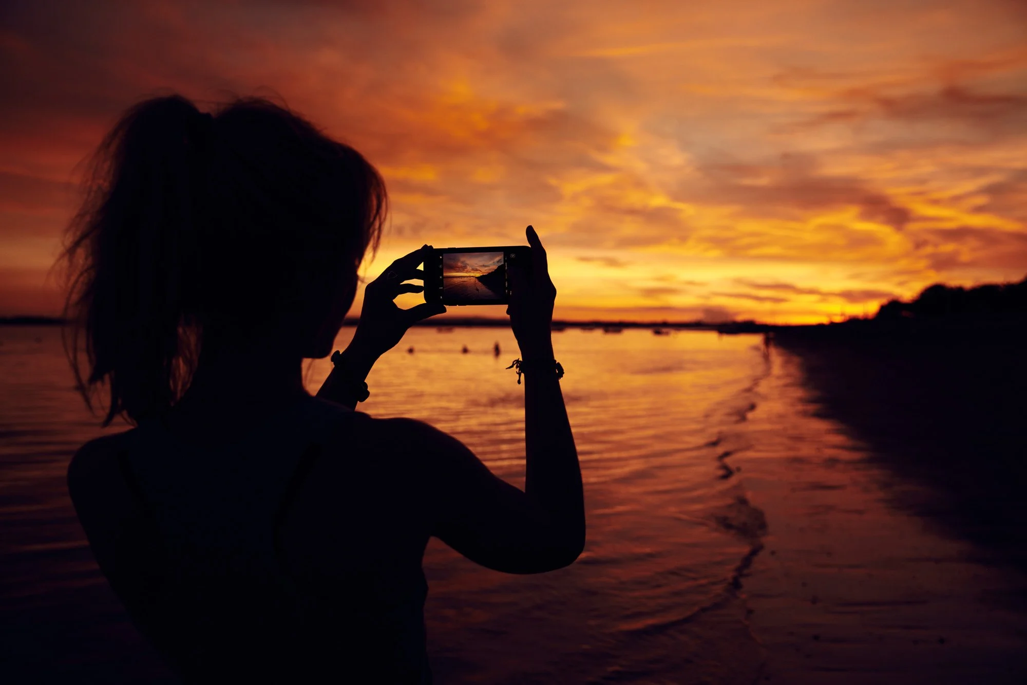 Silhouette of a woman taking a photo of a sunset over the ocean with her phone in Bali Indonesia