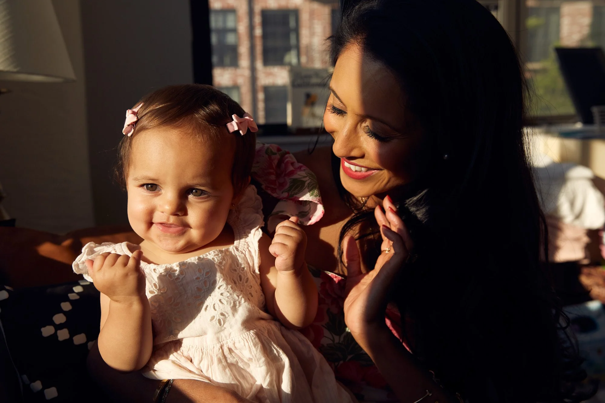 A woman with dark hair and a young girl with light brown hair and pink bows are smiling at each other near a window with sunlight illuminating their faces.