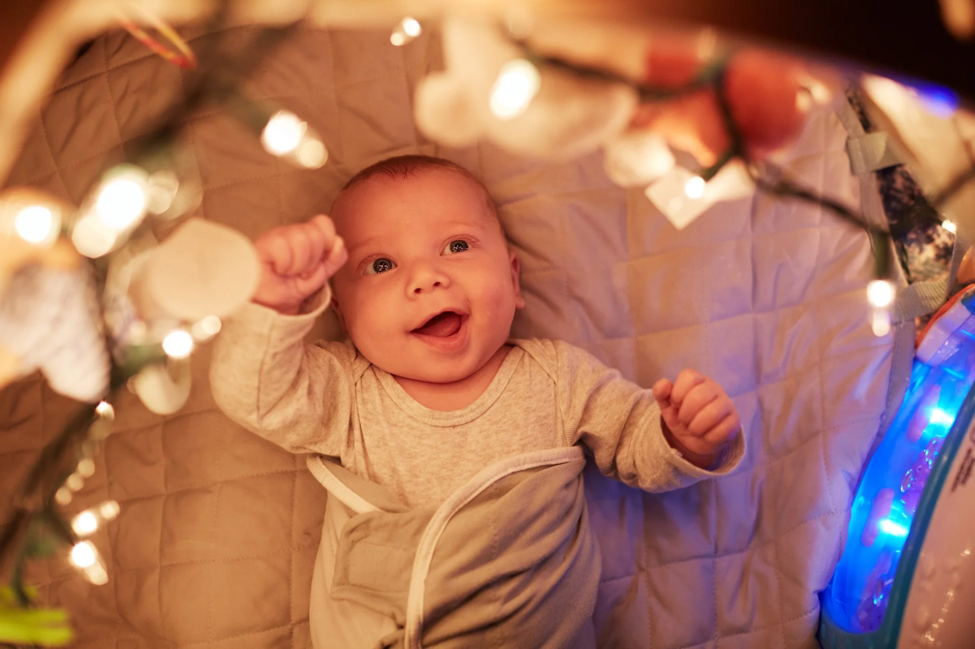 A smiling baby looking up inside a crib decorated with Christmas lights and ornaments.