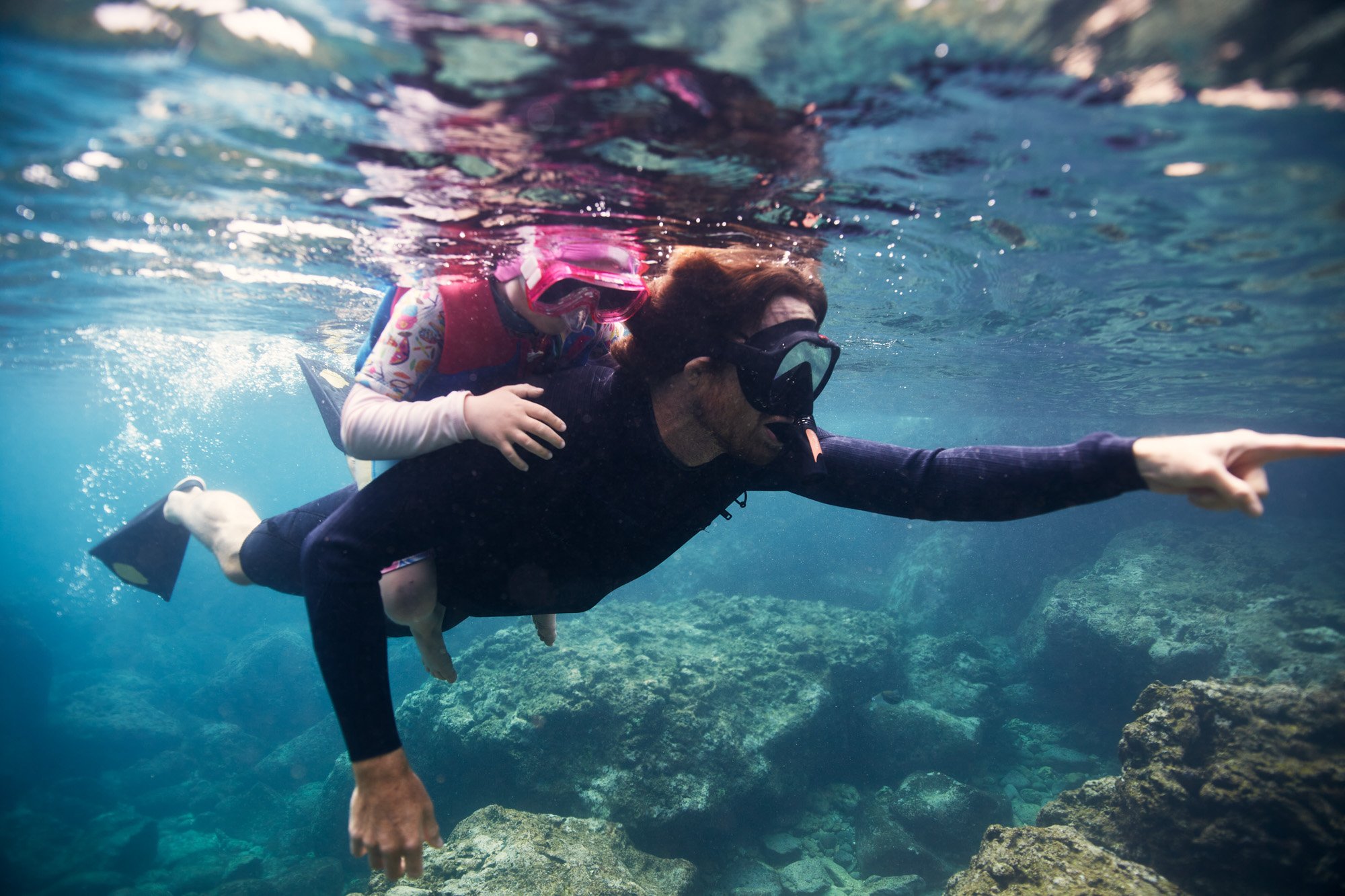 A father and young daughter, both wearing snorkeling gear, swimming underwater near rocks and coral. Sharks Cove Oahu Hawaii