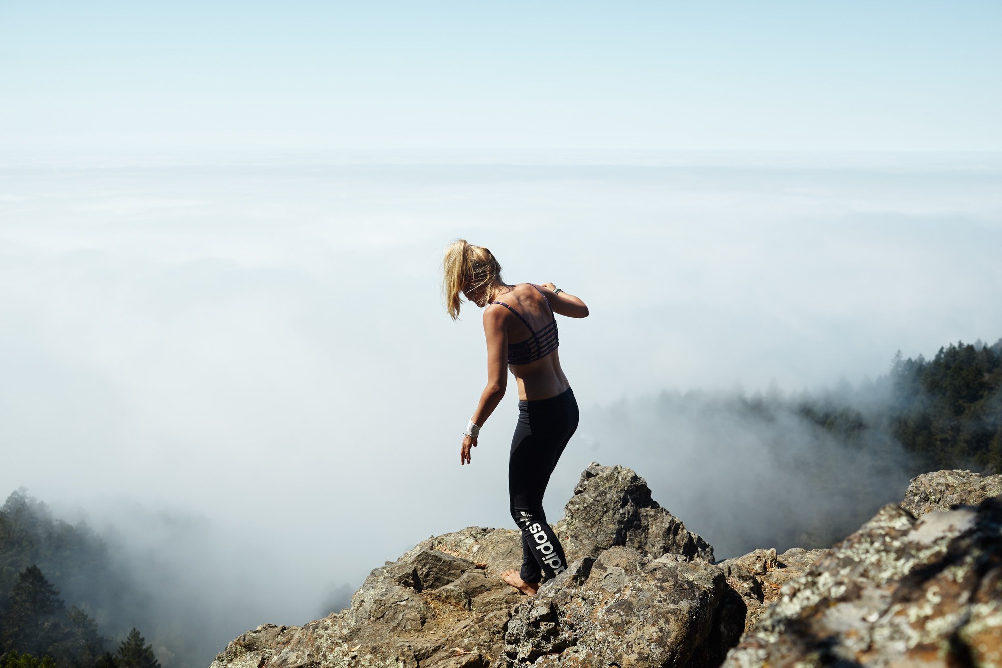 A woman standing on rocky terrain at a mountain overlook near Mount Tamalpais in Marin County California, surrounded by clouds and fog, with forested hills in the distance, wearing athletic clothing.
