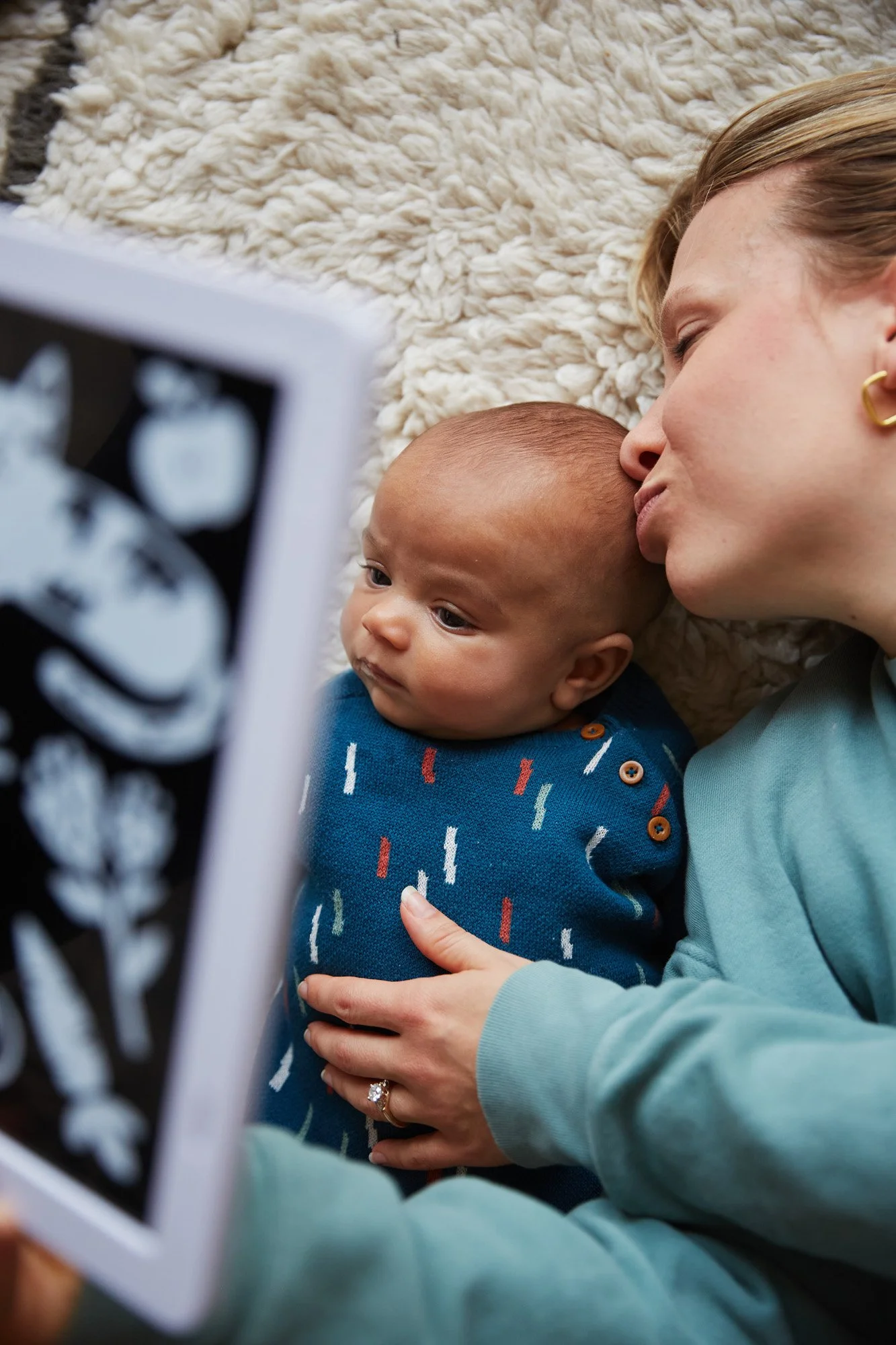 A woman and a baby lying on a soft, cream-colored shaggy rug, looking at an ultrasound picture. The woman is leaning in to kiss the baby's head, holding the baby gently with one hand that has a ring on her finger. The baby is wearing a blue shirt wit