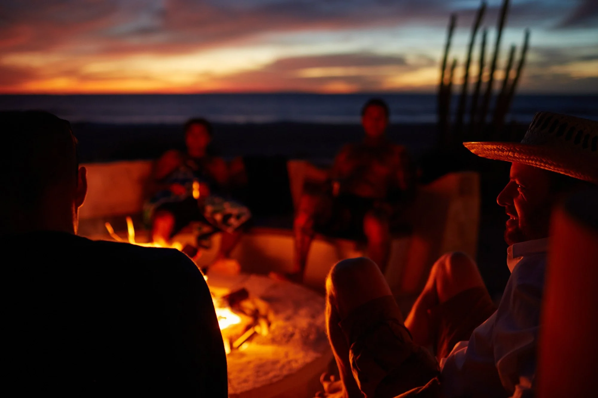 People sitting around a campfire on the beach at sunset, some with drinks, enjoying a relaxed evening outdoors near Todos Santos in Baja California Mexico