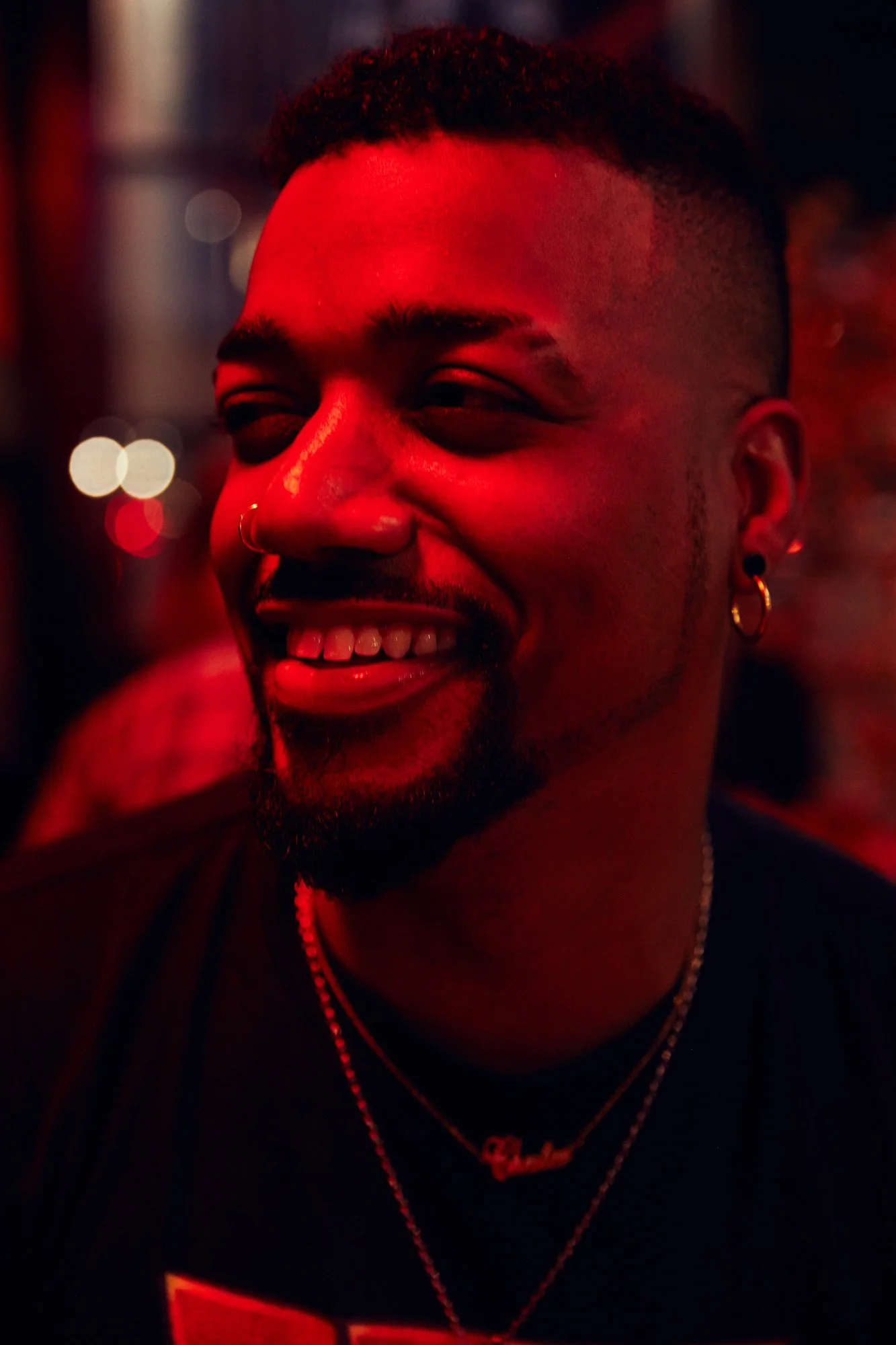 A smiling man with a beard and earrings, illuminated by red lighting, wearing necklaces, in a lively environment in Manhattan New York City