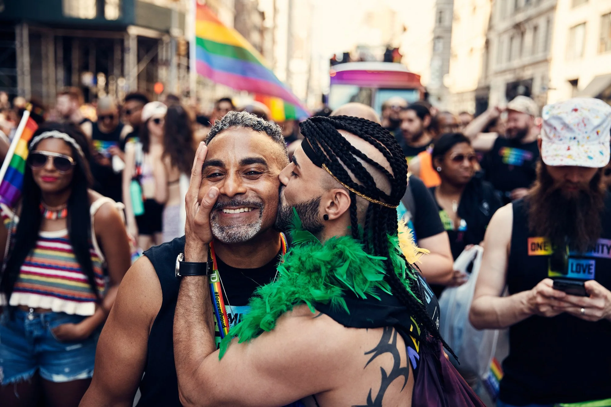 Two men sharing a kiss at NYC pride parade, one smiling and the other about to kiss him, with a crowd of diverse people and rainbow flags in the background.