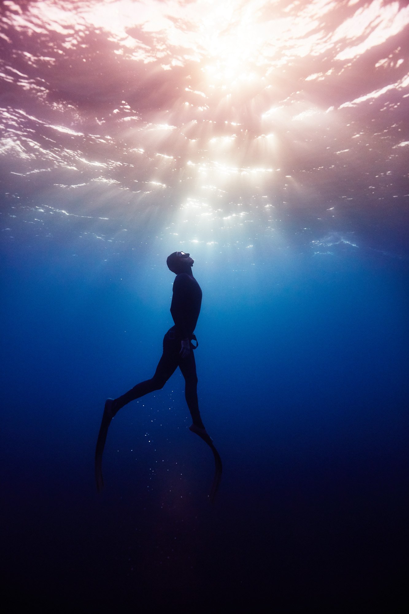 A person underwater using fins, looking up towards the sunlight streaming through the water surface.