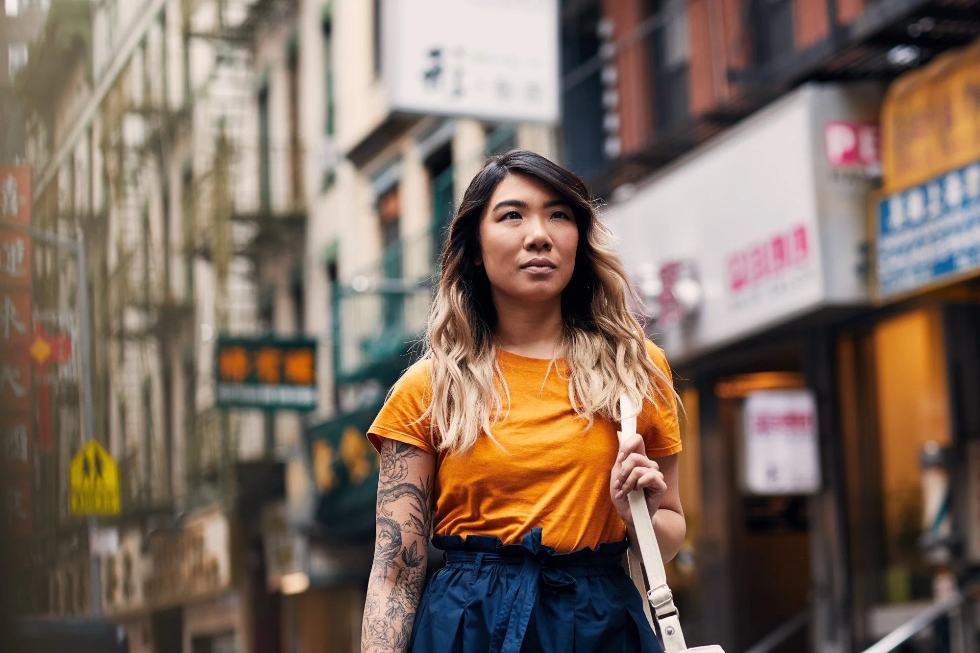 Young woman Carol Lee with tattoos on her arm, wearing an orange t-shirt and navy high-waisted pants, walking on a city street in Chinatown Manhattan New York City
