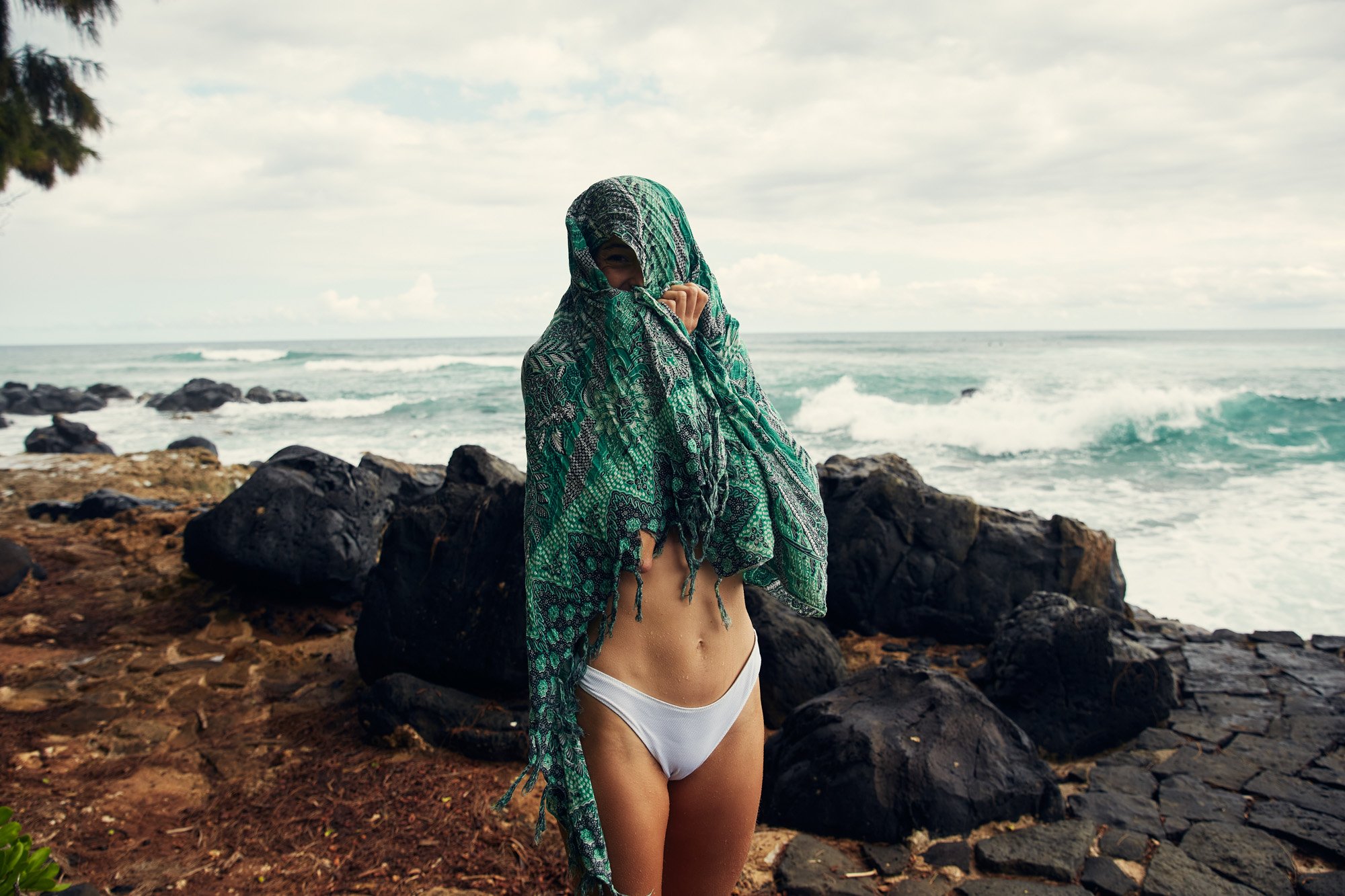 Young woman in a white bikini and green patterned cover-up shawl at a rocky beach with ocean waves and cloudy sky, on the south shore of Oahu Hawaii