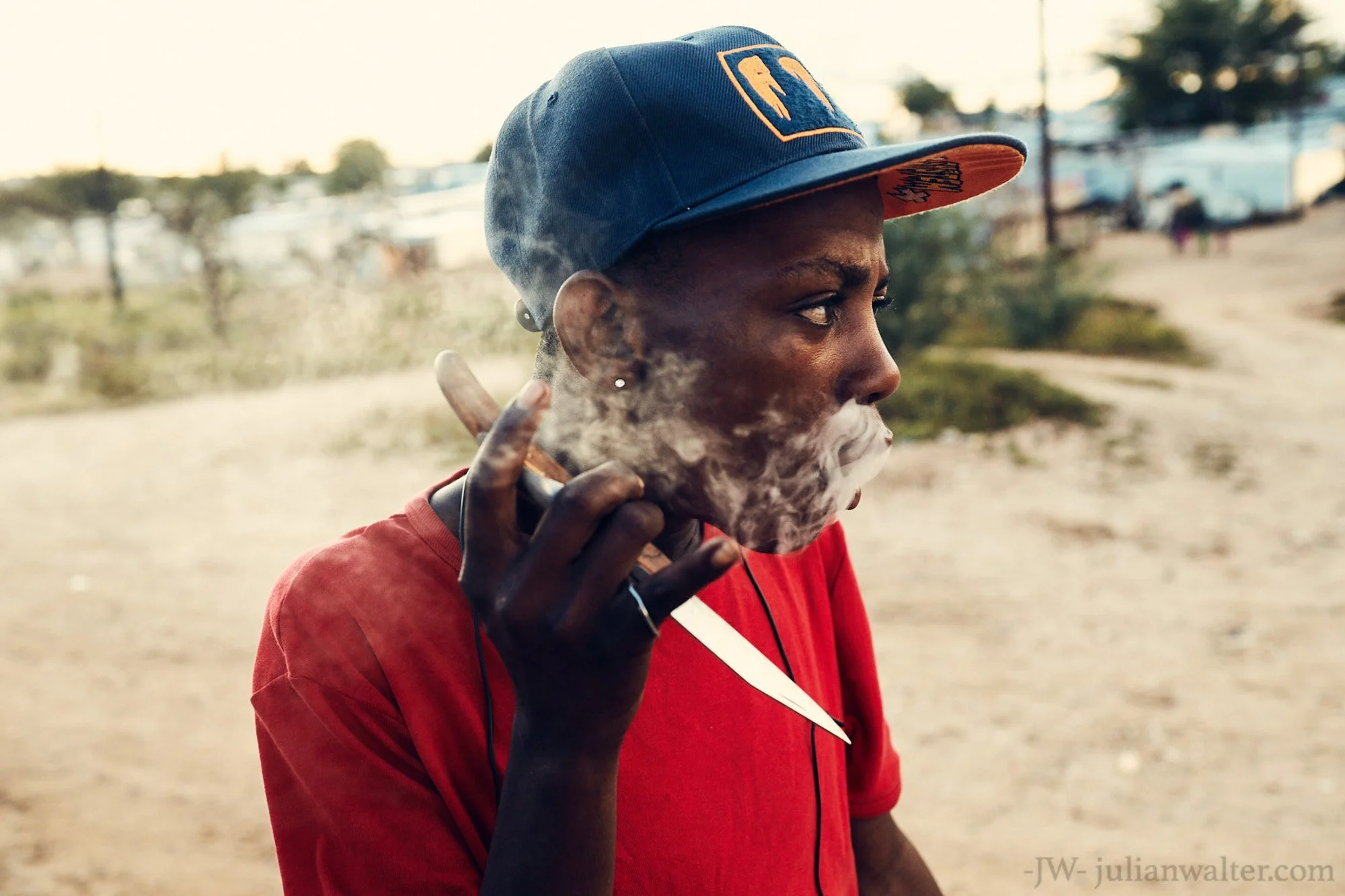 A young man wearing a blue baseball cap and red shirt smokes a cigarette outdoors in the townships of Windhoek Namibia, with smoke coming out of his mouth wrapping around his face and a blurred background of tents and trees.