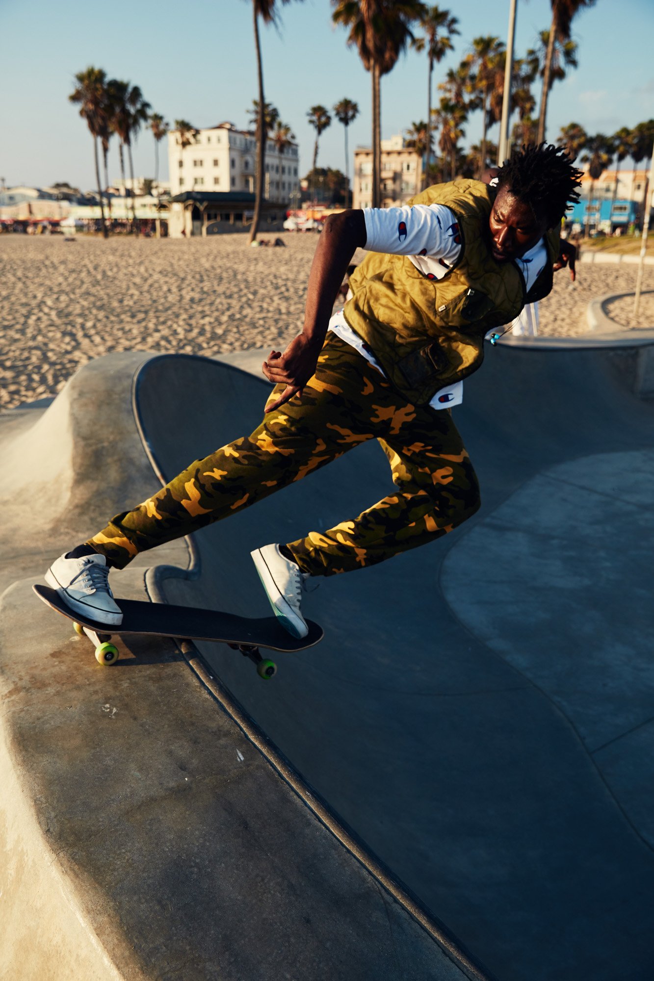 Skateboarder performing a trick at Venice Beach skate park near the beach with palm trees and buildings in the background.