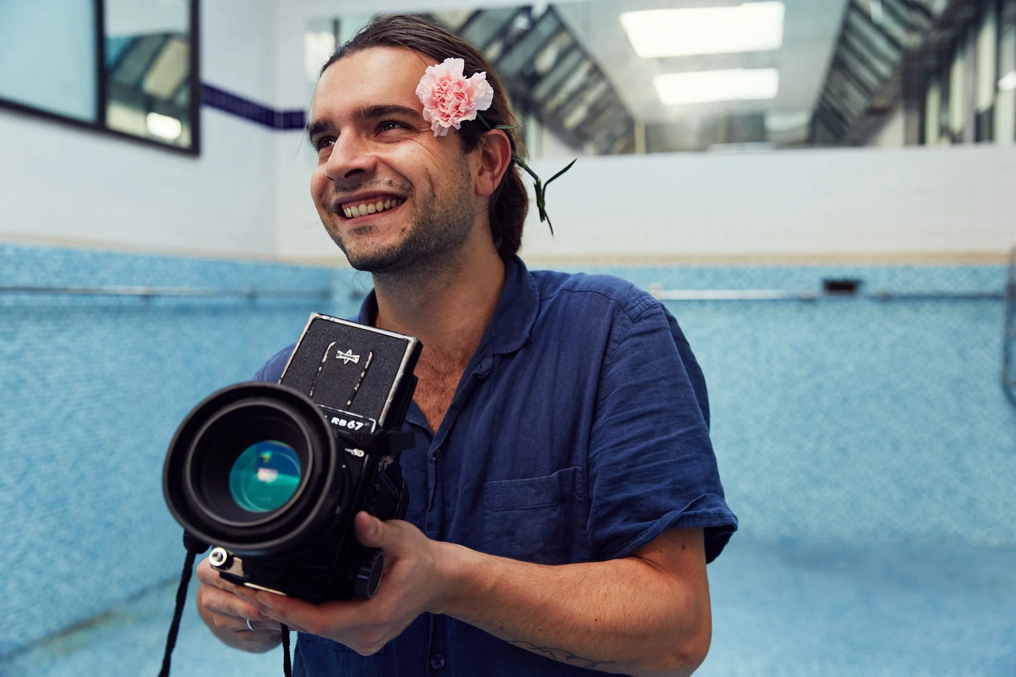 Photographer Henri Kisielewski with a pink flower behind his ear, holding a medium format film camera, smiling, in an empty swimming pool