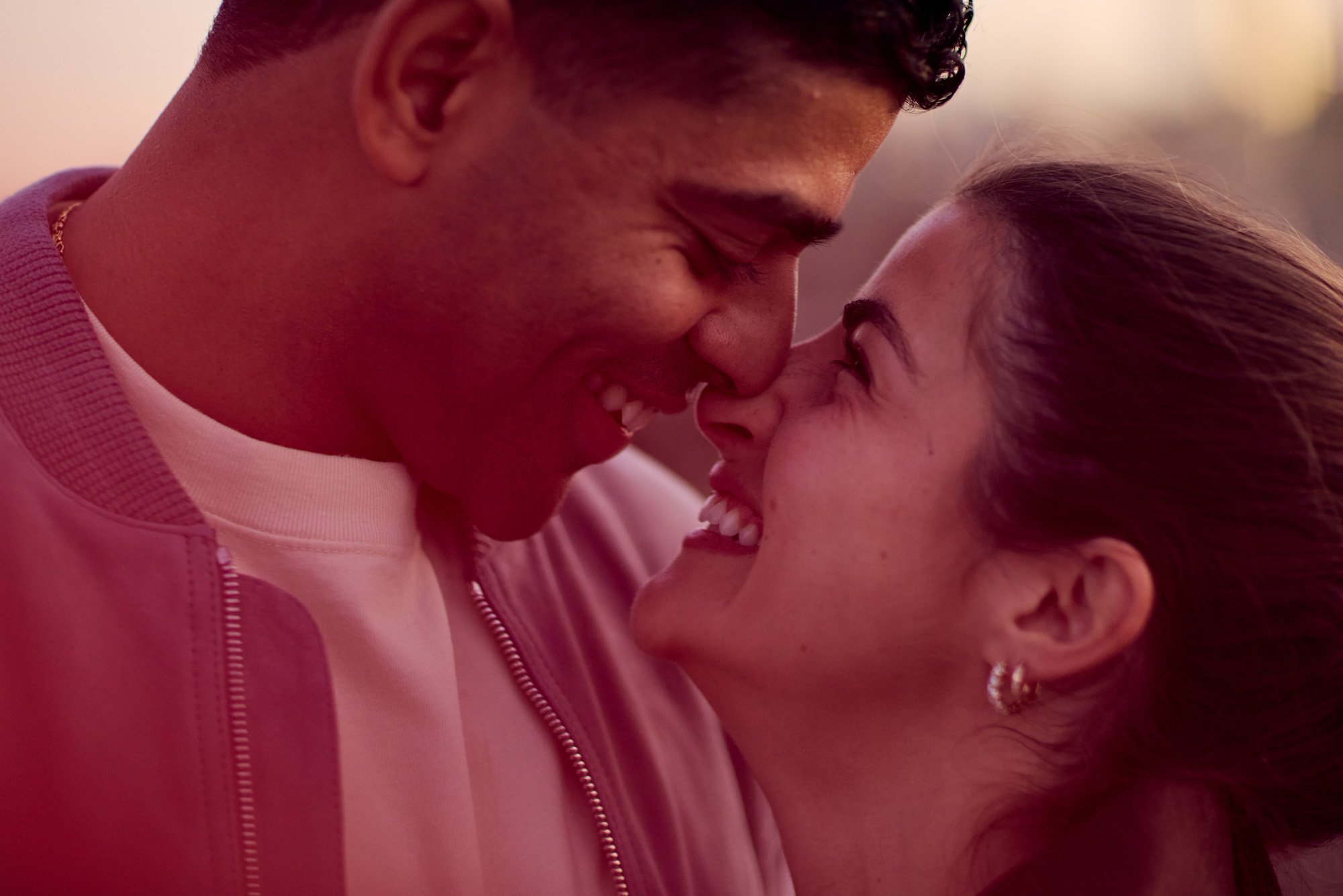 A man and woman are close together, smiling and touching noses, with eyes closed, in a tender and affectionate moment.