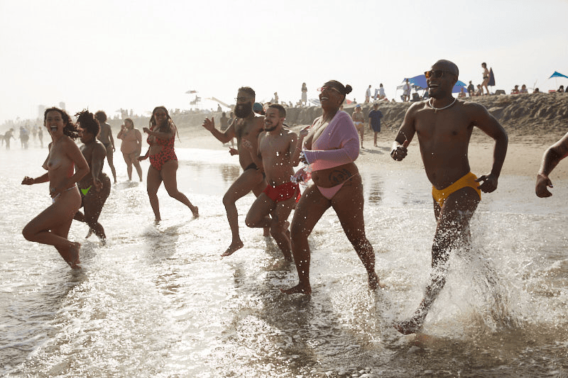 Group of diverse people smiling and playing in the ocean waves at the beach with umbrellas and other beachgoers in the background. Pride Jacob Riis Beach New York City
