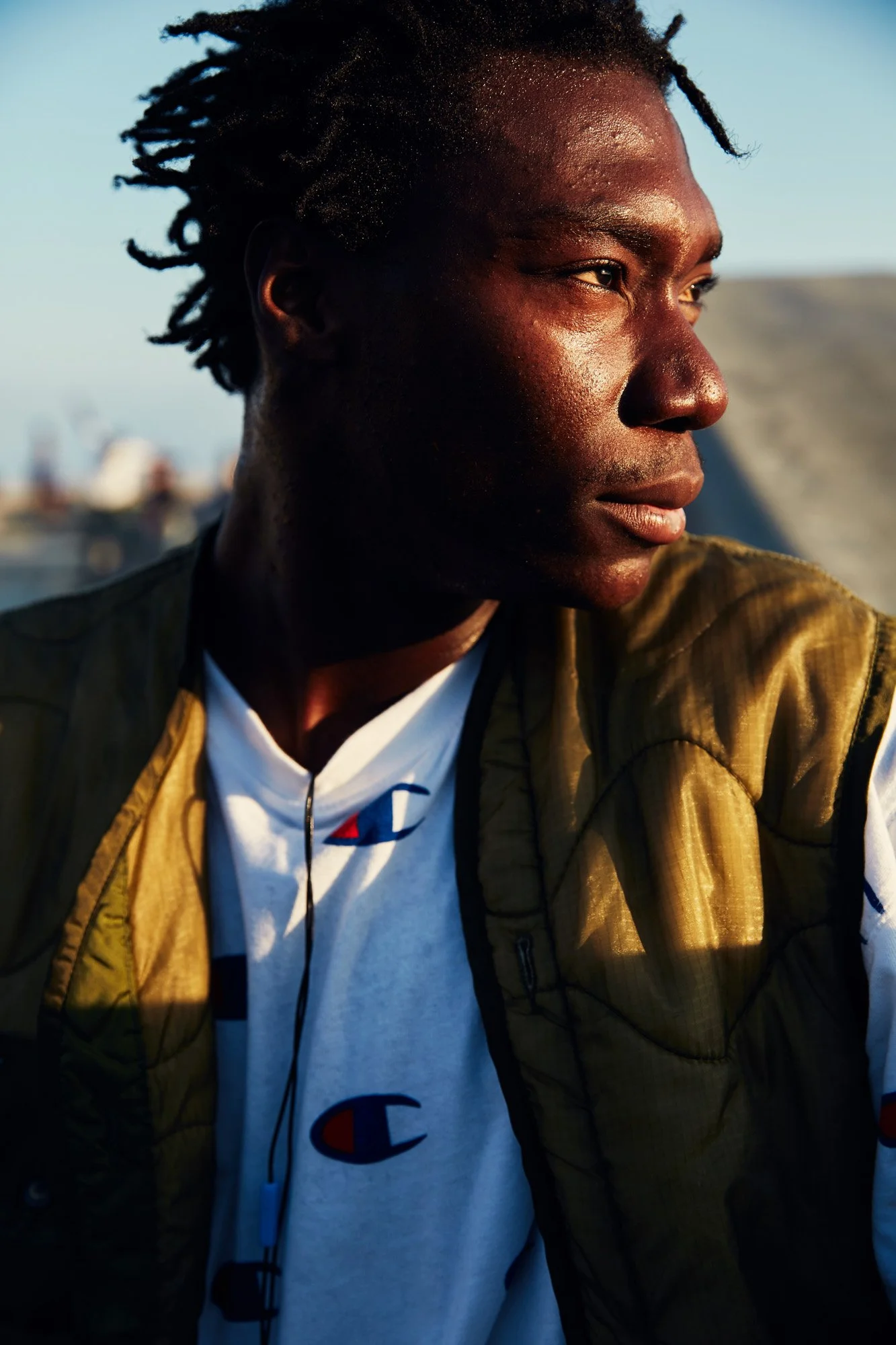 A young man with dark skin and short dreadlocks gazes to the right in a side profile shot. He is wearing a white Champion t-shirt and a brown vest, and is outdoors with a blurred urban background during daylight.