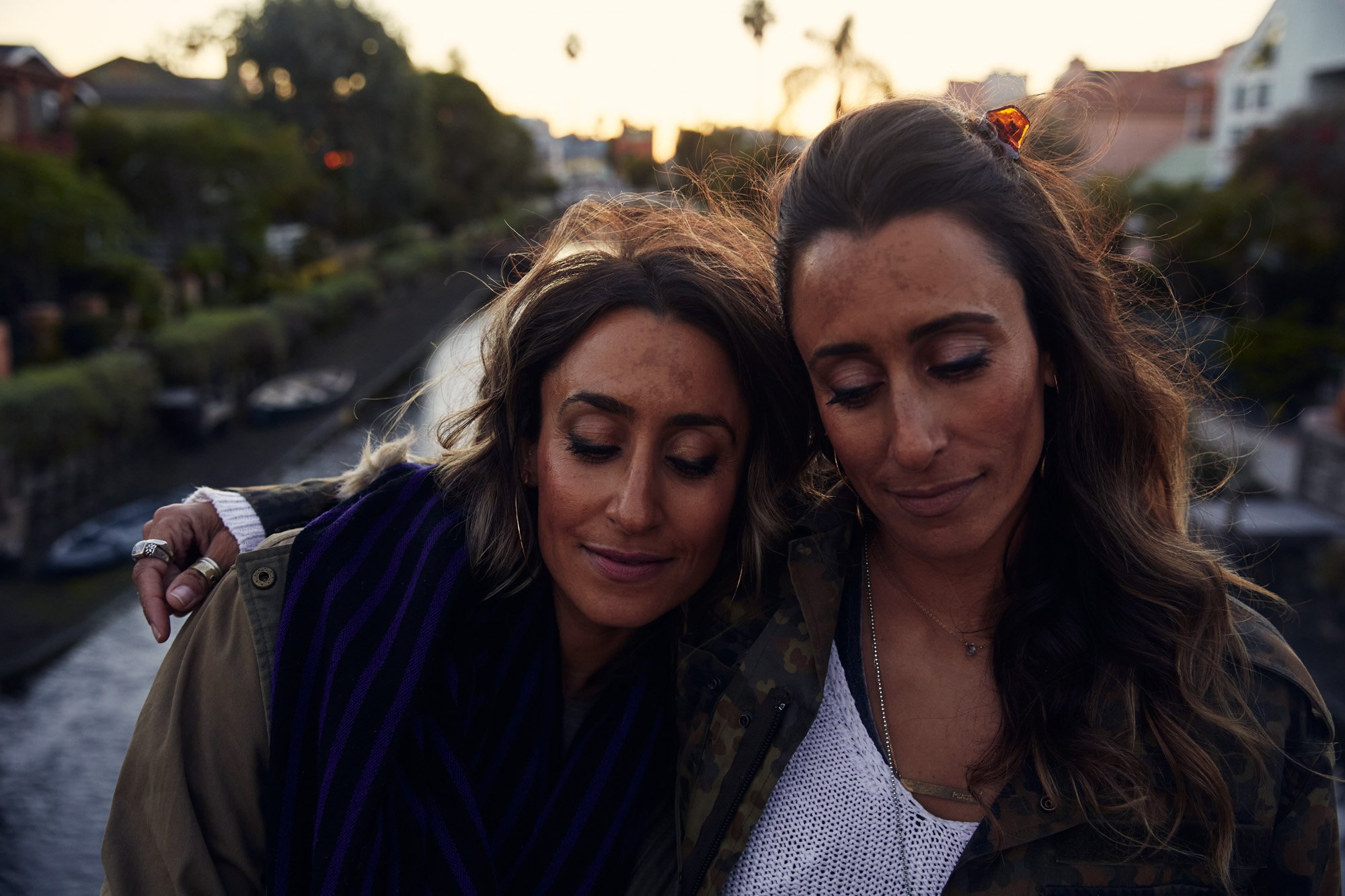 Two identical twins with closed eyes sharing an embrace outdoors during sunset with trees and houses in the background.