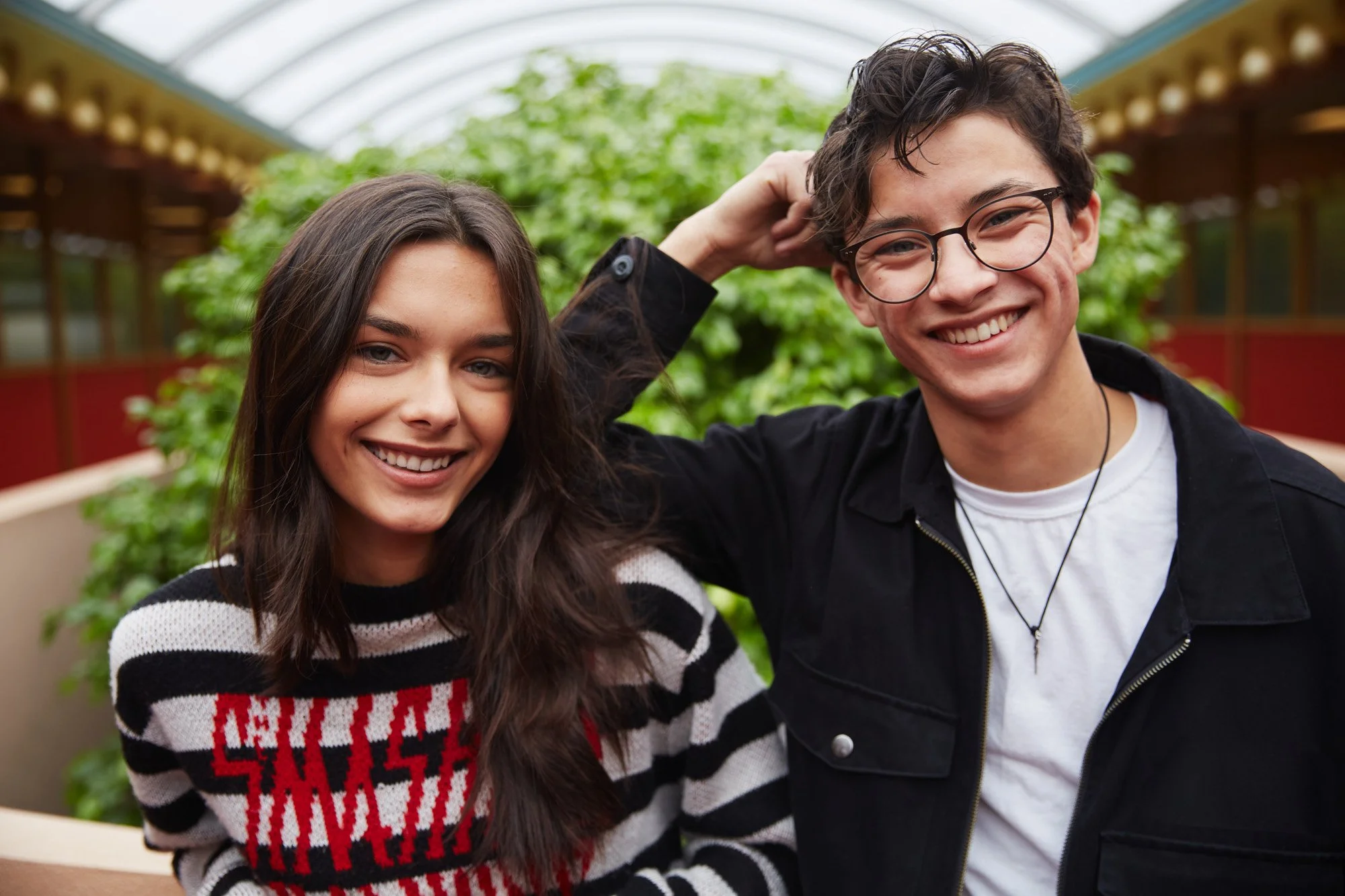 Smiling young woman Violet Lux and man posing together in a greenhouse with plants in the background.