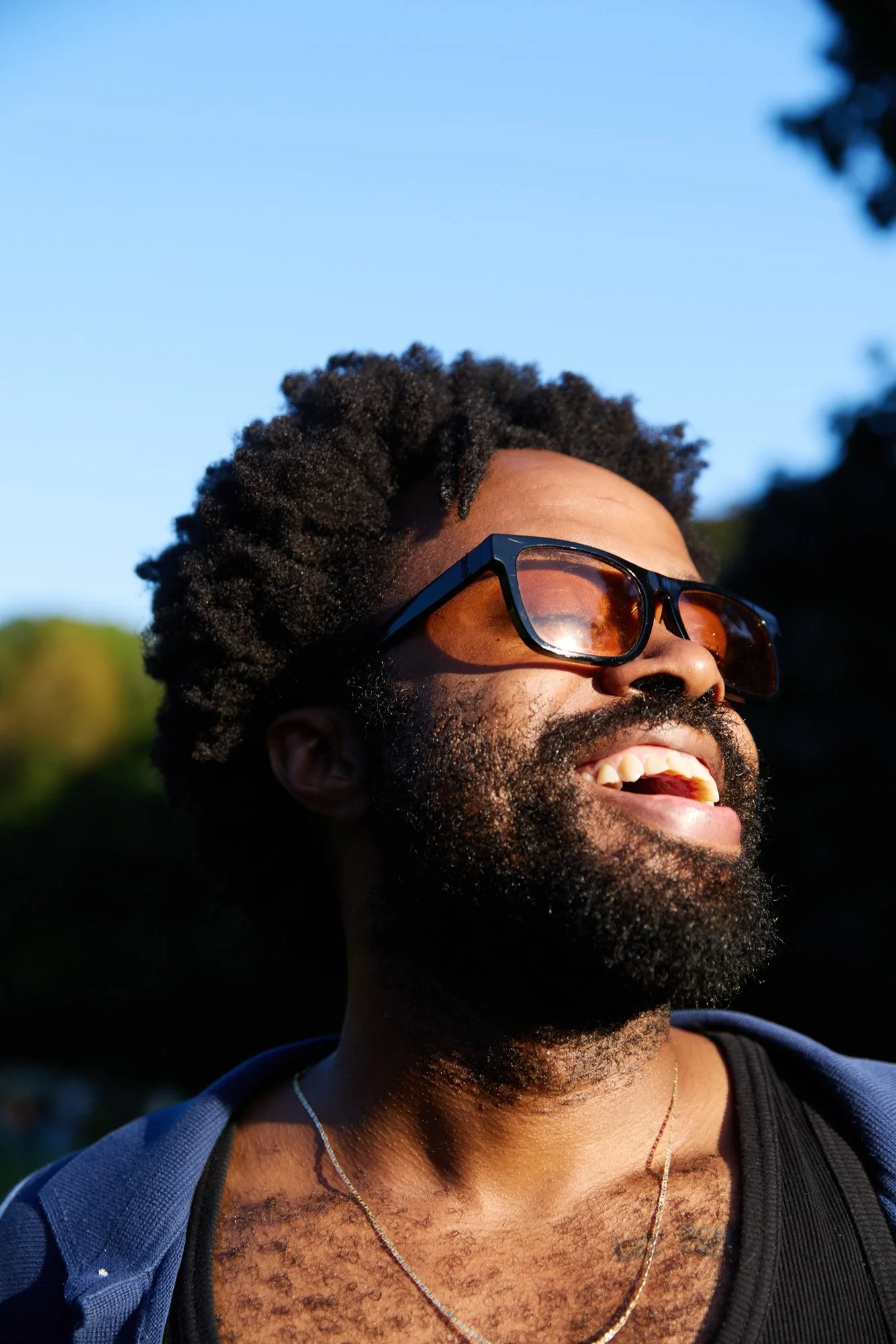 A joyful Black man with curly hair, beard, and sunglasses smiling outdoors in sunlight, wearing a black shirt and a gold chain.