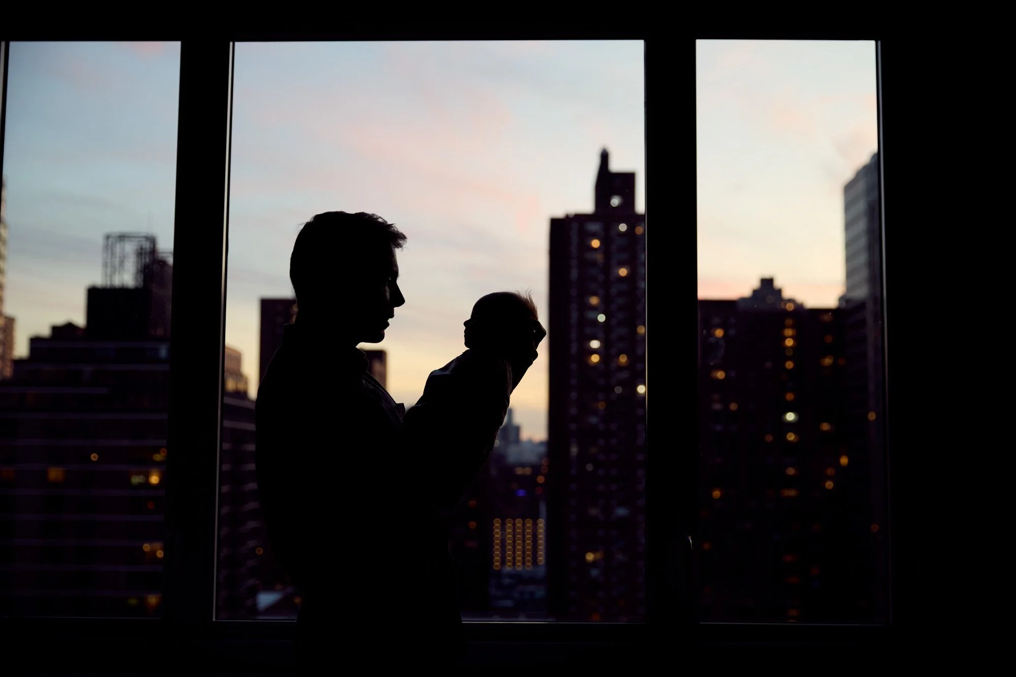 Silhouetted man holding a baby in front of large windows overlooking the New York City skyline in the evening after sunset.
