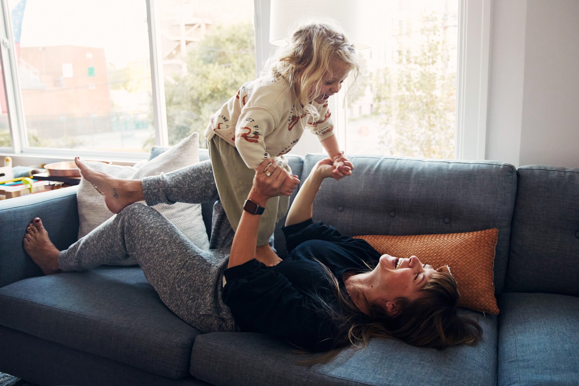 A mother and her toddler daughter playfully wrestling on a gray couch with a window in the background.
