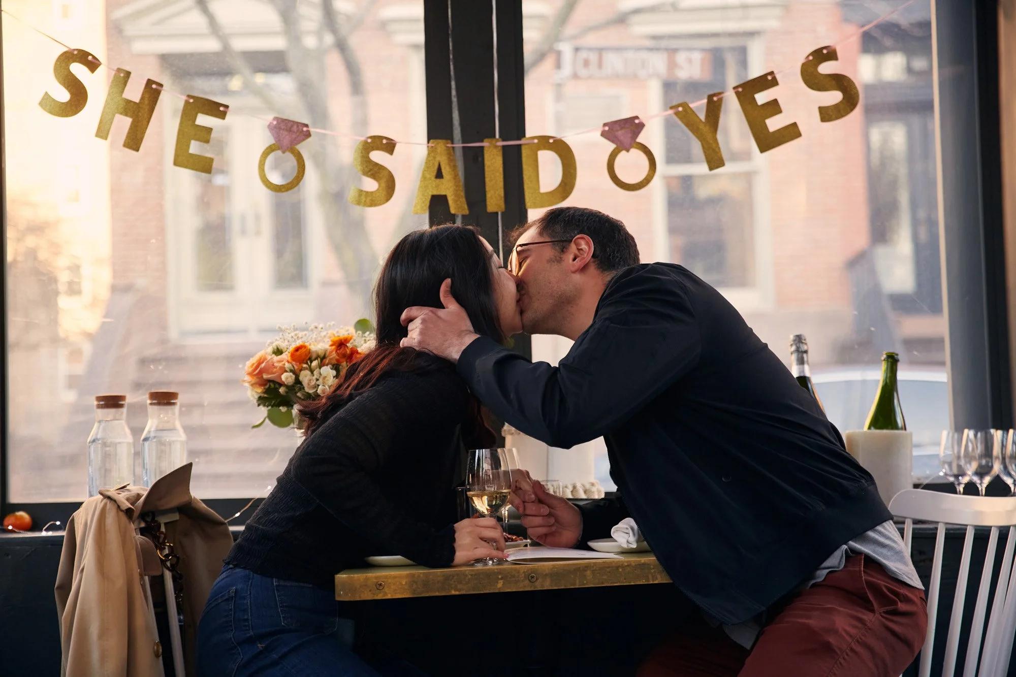 A man and woman sharing a kiss at a table in a restaurant with a sign that says "She said yes" next to them