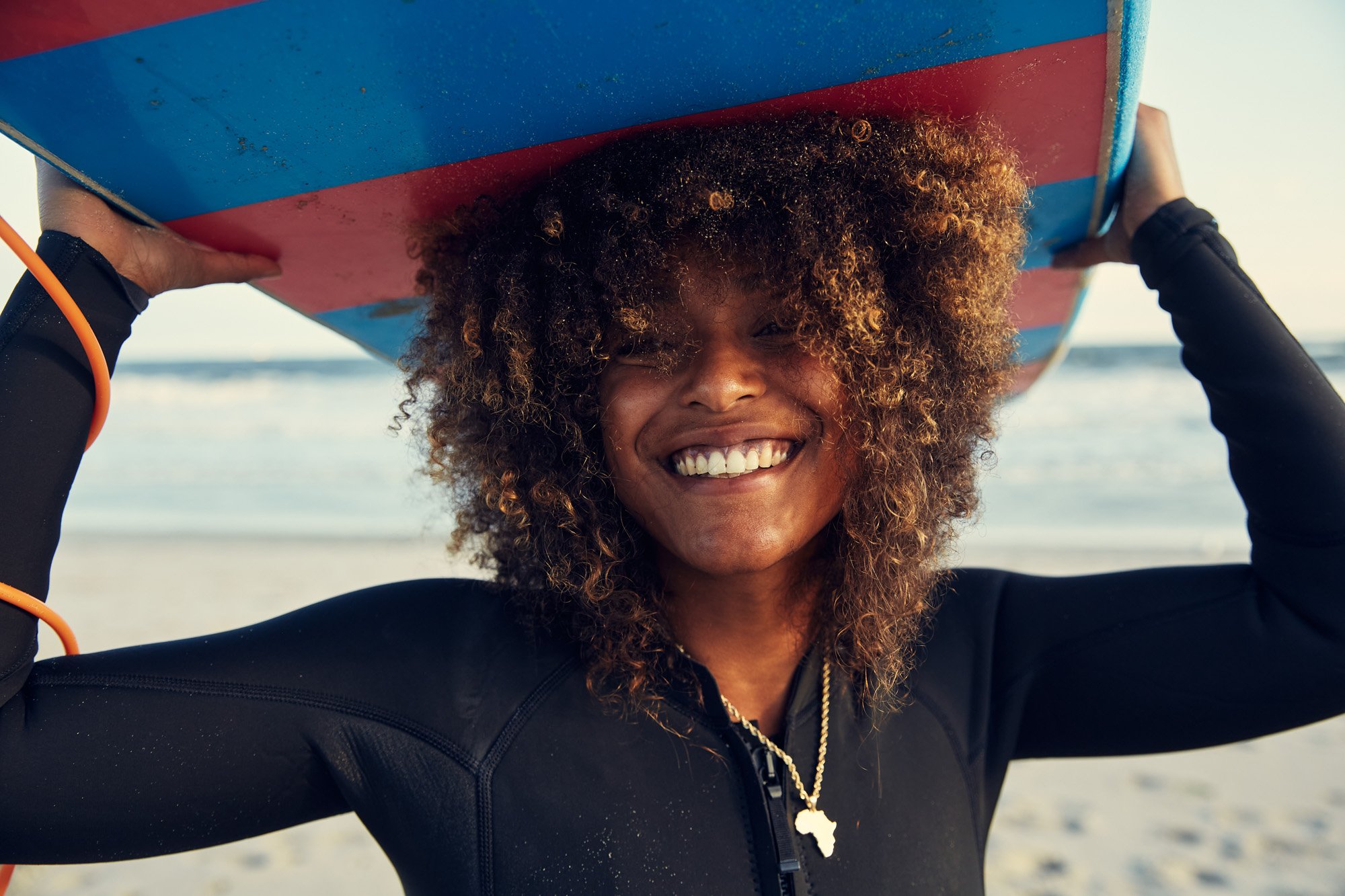Smiling woman holding a surfboard on her head at the beach, wearing a black wetsuit and a necklace with an Africa-shaped pendant at Rockaway Beach New York City.