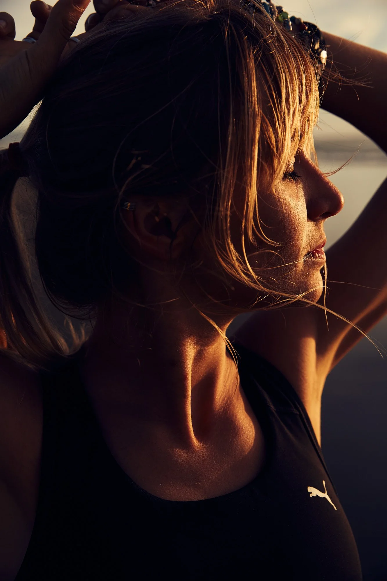 A woman with closed eyes, golden hair, and a black Puma sports top, standing outdoors at sunset at Ocean Beach San Francisco with her arms raised behind her head.