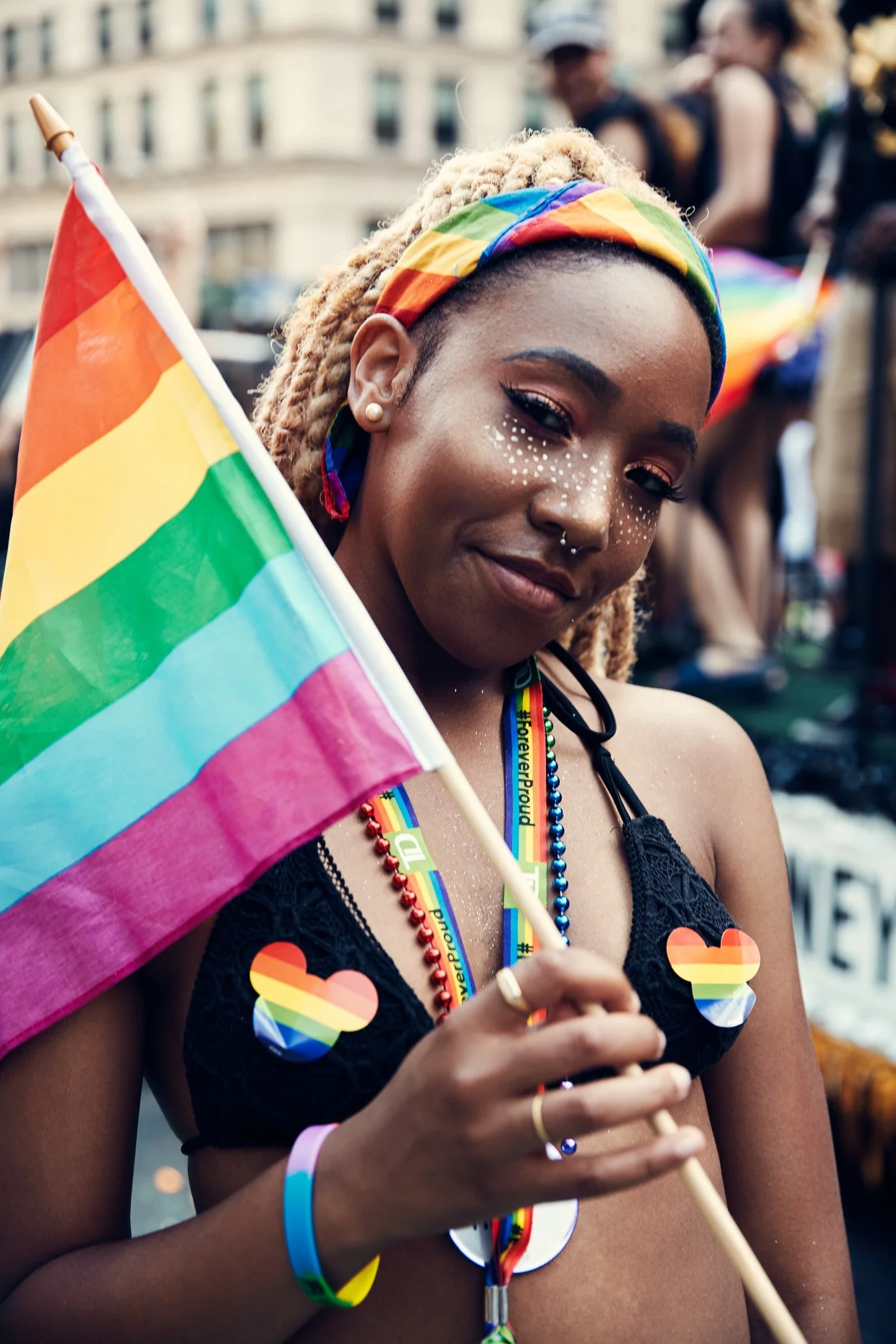 A woman at NYC pride parade holding a rainbow flag and wearing rainbow-themed accessories, including stickers and bracelets, smiling with face glitter and a colorful bandana.