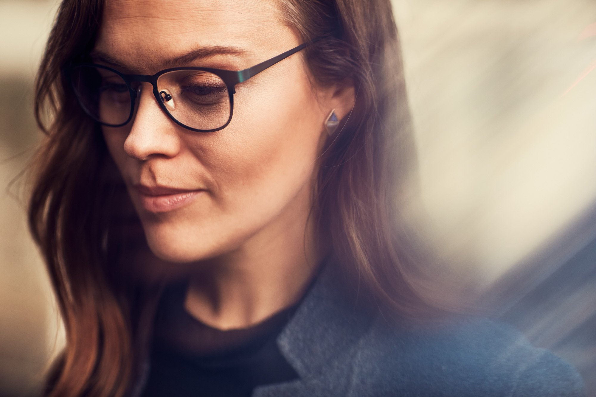 Close-up of a woman with brown hair, wearing glasses with black frames and a dark sweater, looking down.