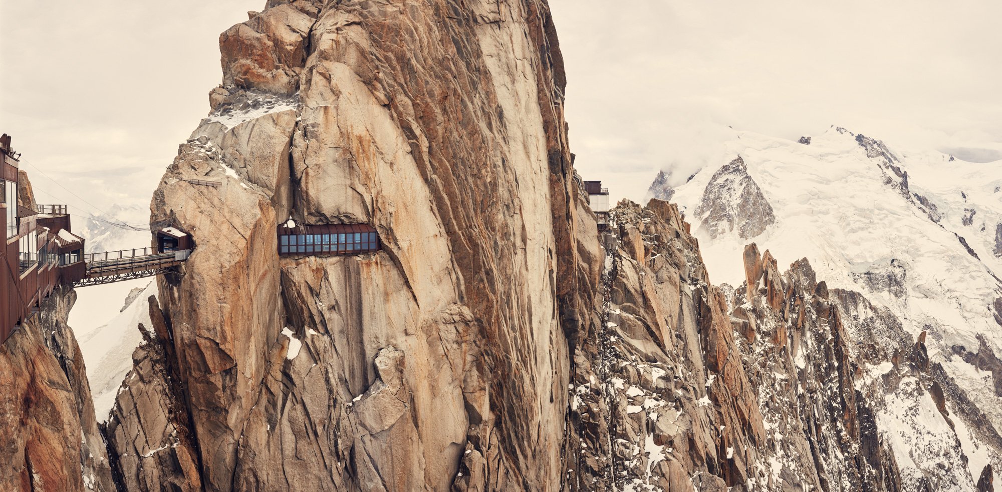A rock mountain with a glass observation deck built into the side of a large rock face, snow-covered peaks in the background. Aiguille du Midi, Chamonix, France