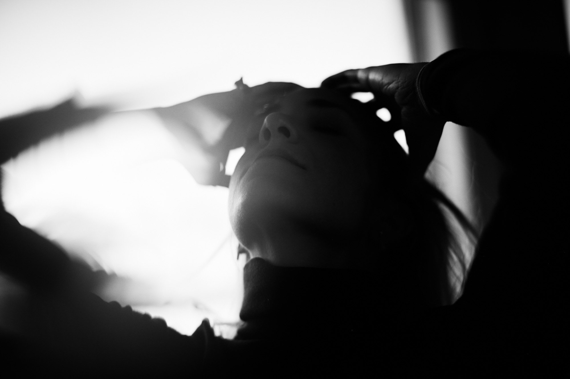 A black and white photo of Sami Freeman with her eyes closed, holding her head with one hand, with a bright light behind her.