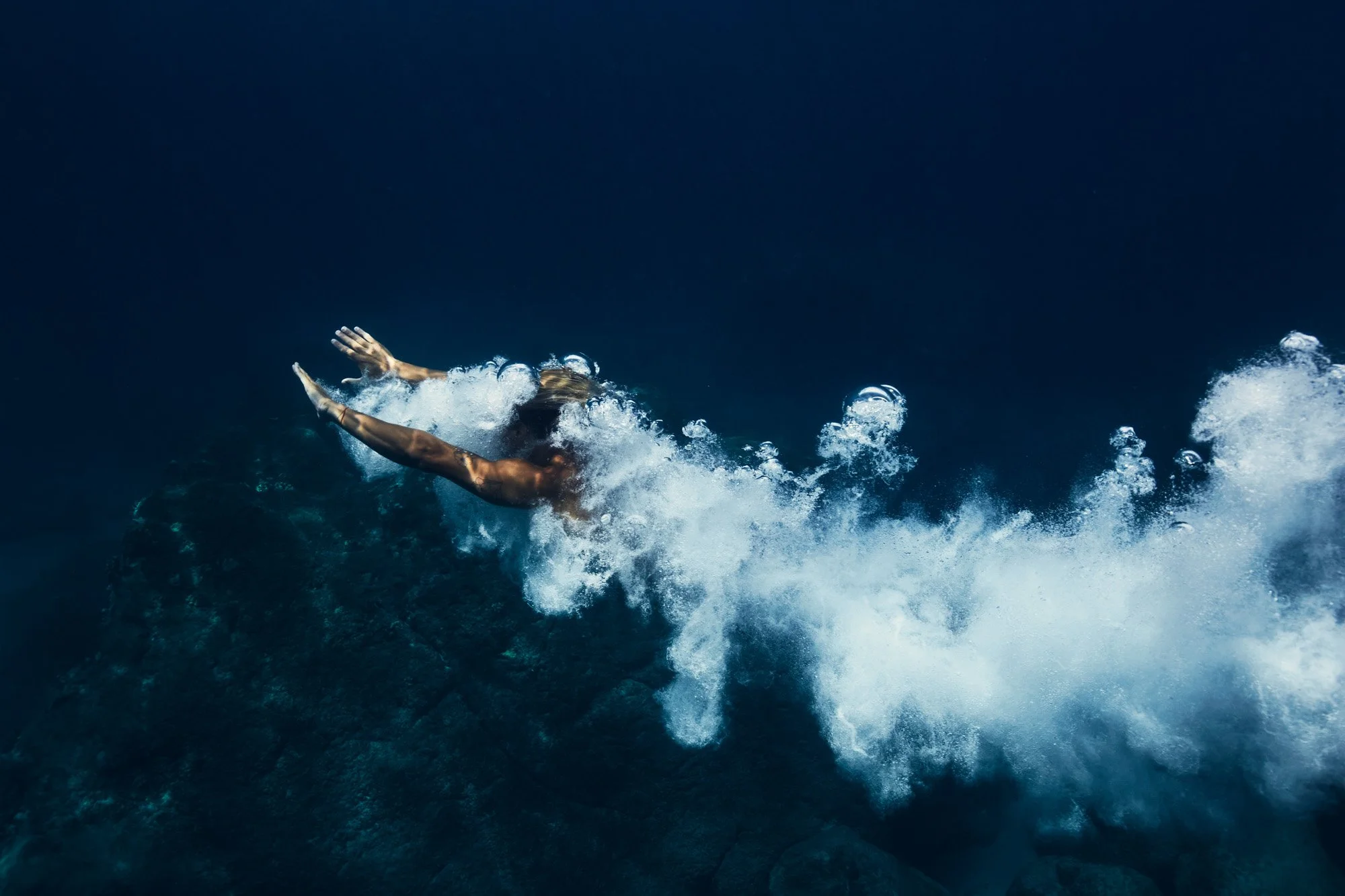 A man diving into a dark blue ocean in Waimea Bay Oahu Hawaii, creating a trail of bubbles.