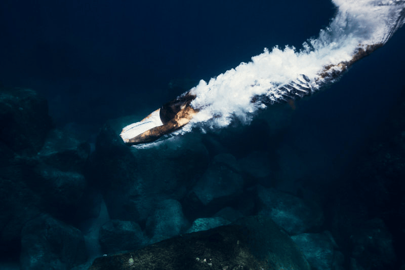 A man just after a dive, swimming underwater over rocks, with a trail of bubbles following behind it.