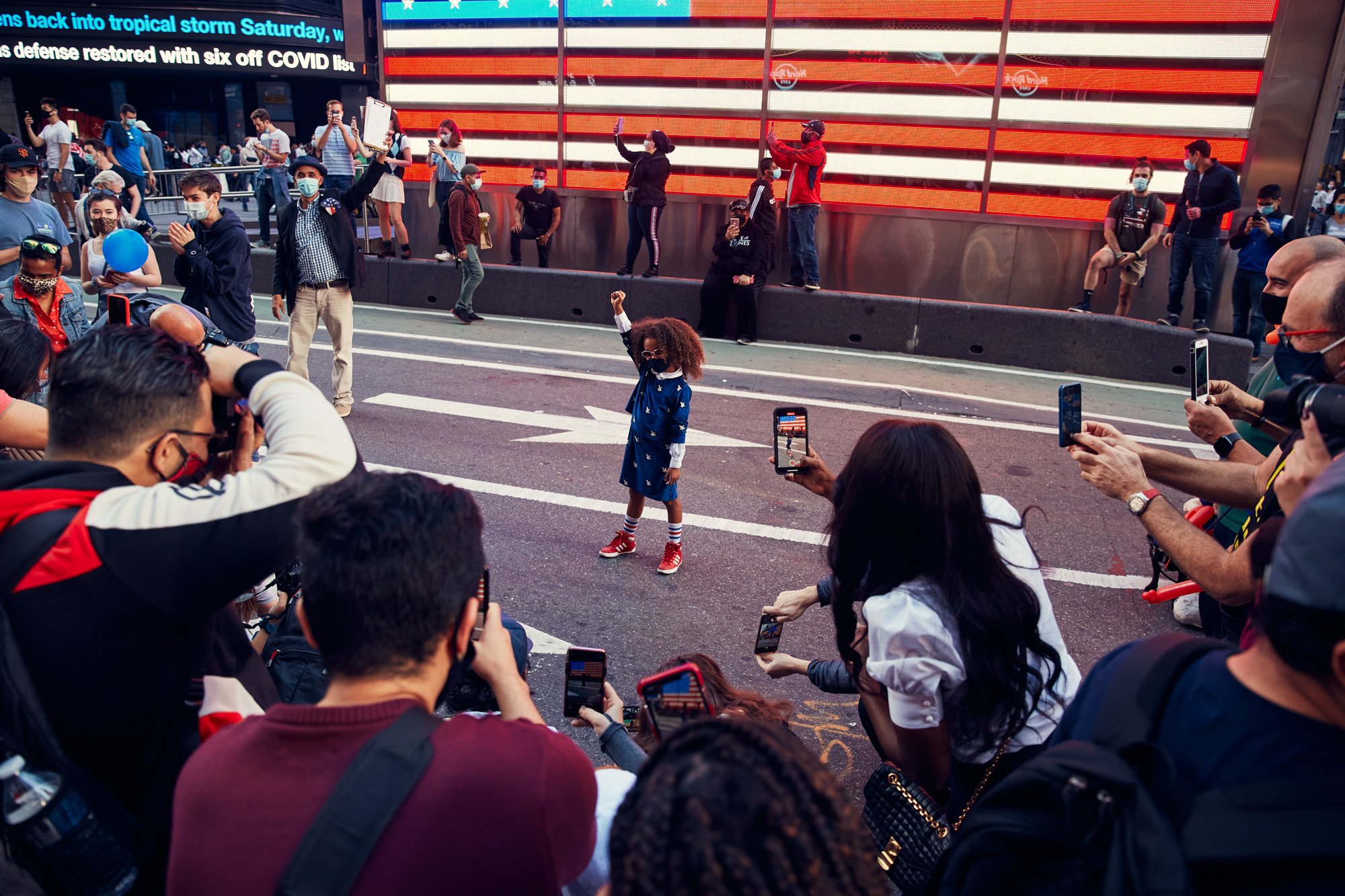 A crowded street scene near Times Square New York City, with people taking photos, a girl dancing in the middle, and some people standing on a raised platform in front of an American flag display. Biden Election Day 