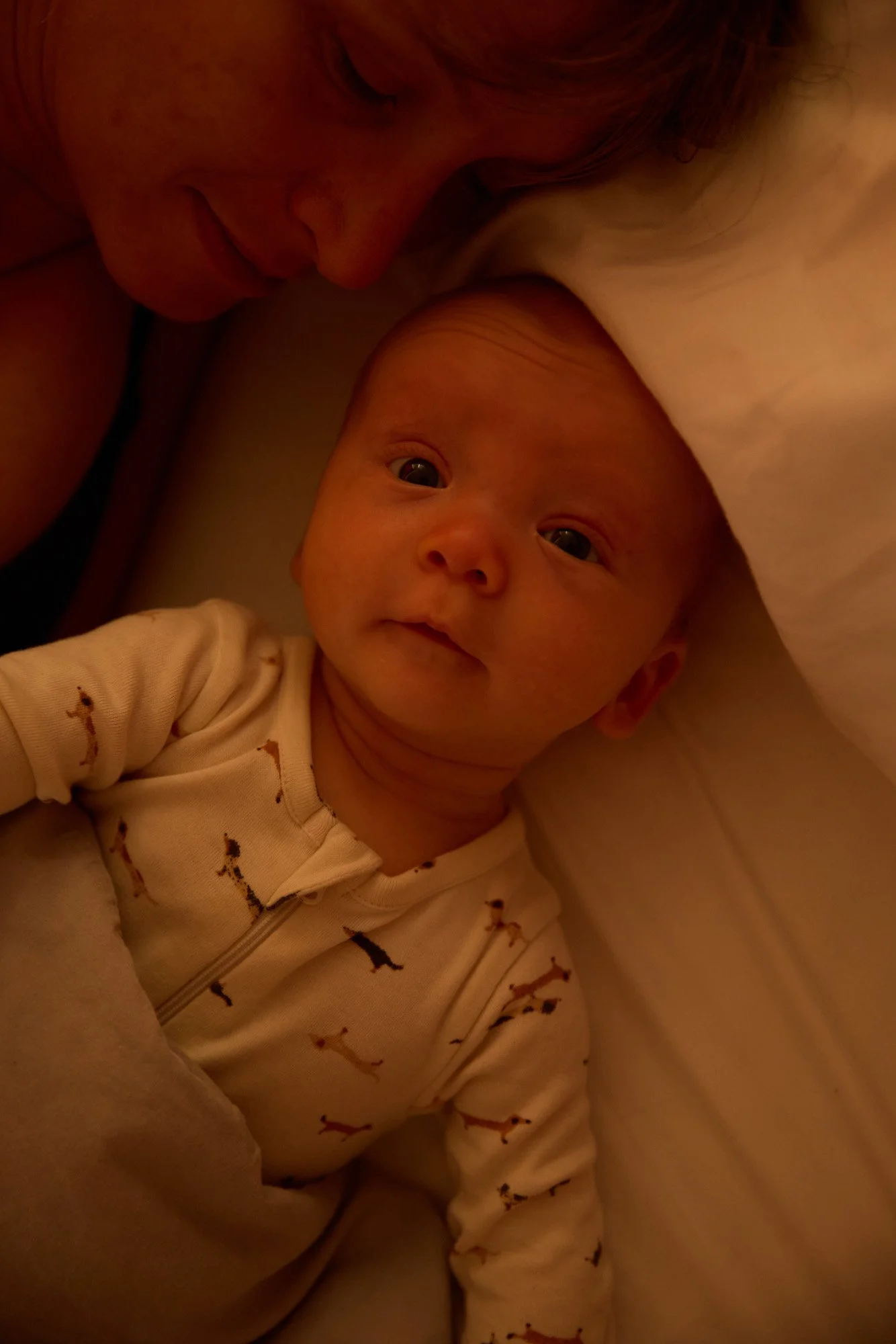 A close-up photo of a baby lying on a bed, with an adult woman leaning over and gently touching the baby's head, both in a dimly lit room.