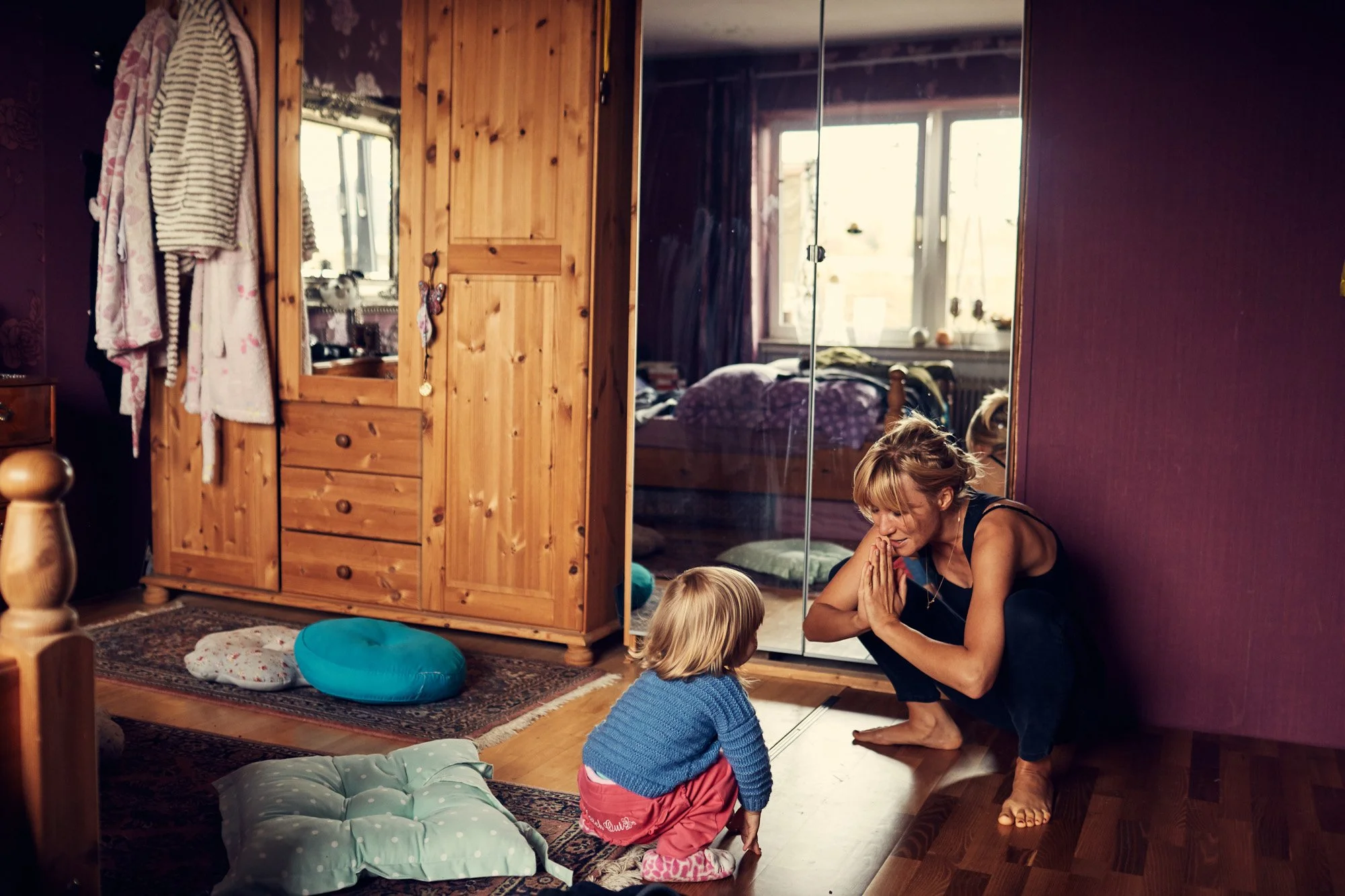 A woman and a small girl are squatting facing each other on the wooden floor in a cozy bedroom. The woman is smiling and has her hands together in a prayer gesture, while the girl is looking at her with a smile. Behind them, there are two cushions on