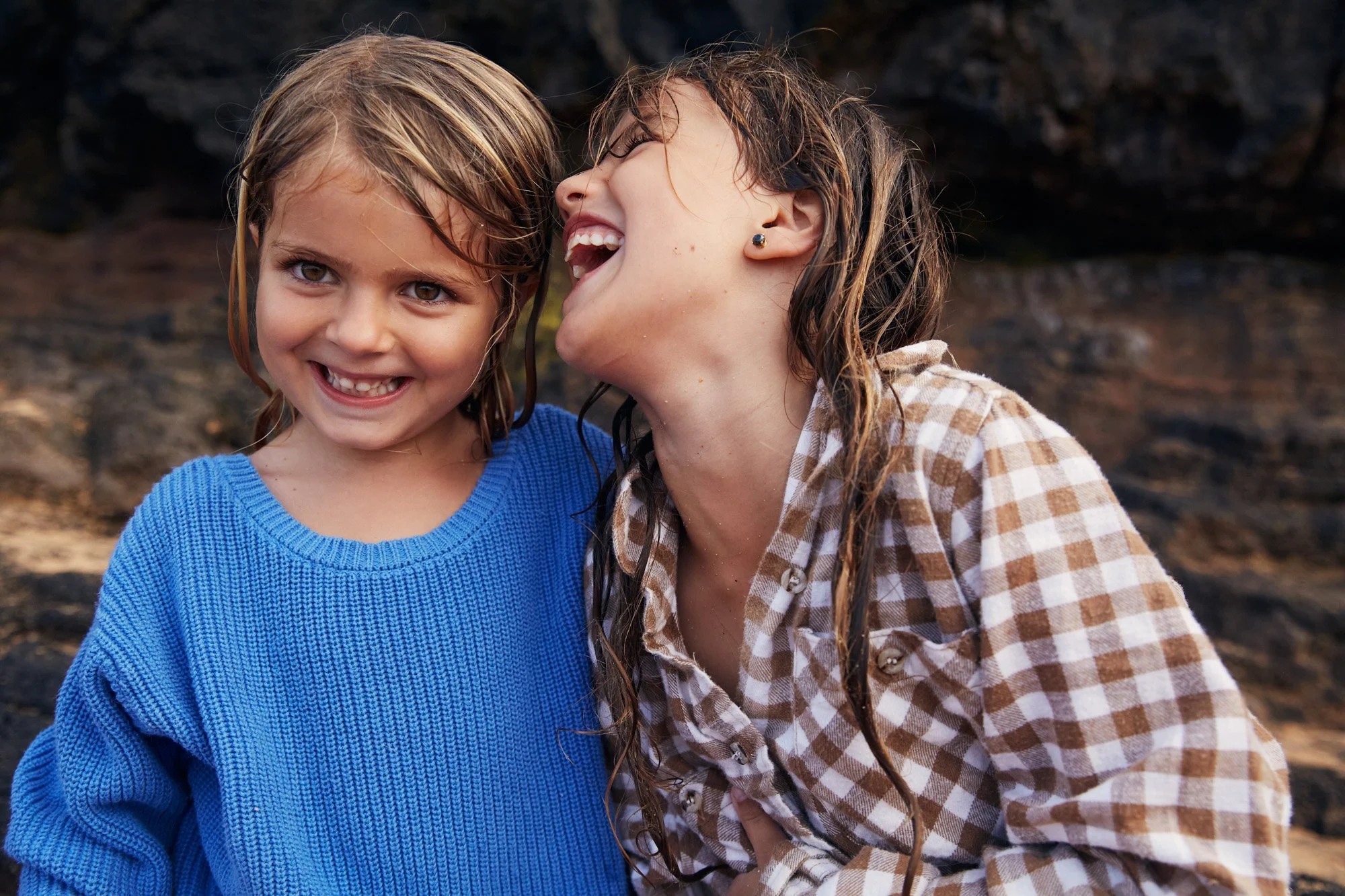 Two young girls smiling and laughing outdoors on a rocky background, one wearing a blue sweater and the other in a checkered shirt.